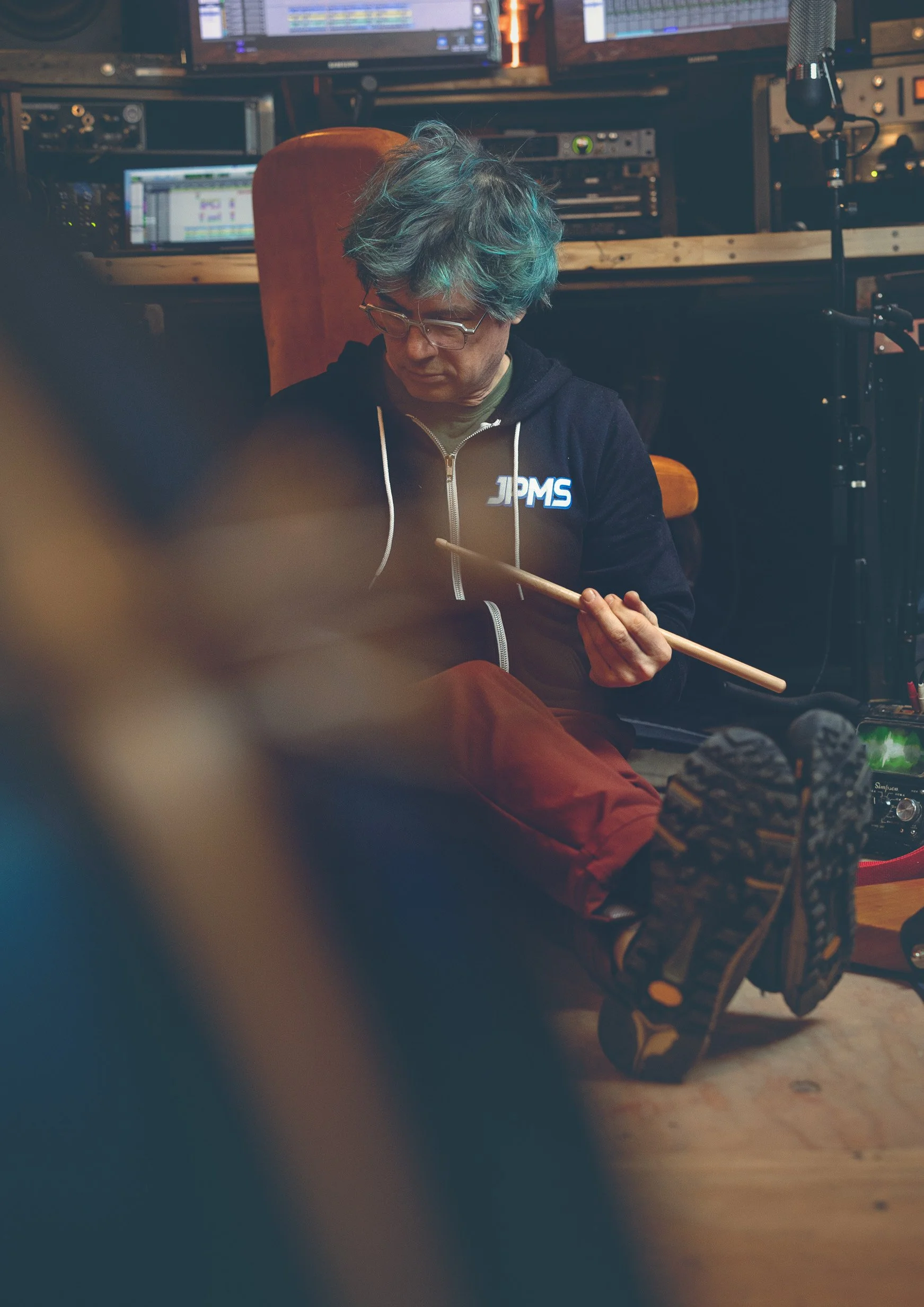 A person with teal and grey hair, wearing glasses and a black hoodie with "JPMS" on it, sitting on the floor in a recording studio, holding a drumstick and looking down.