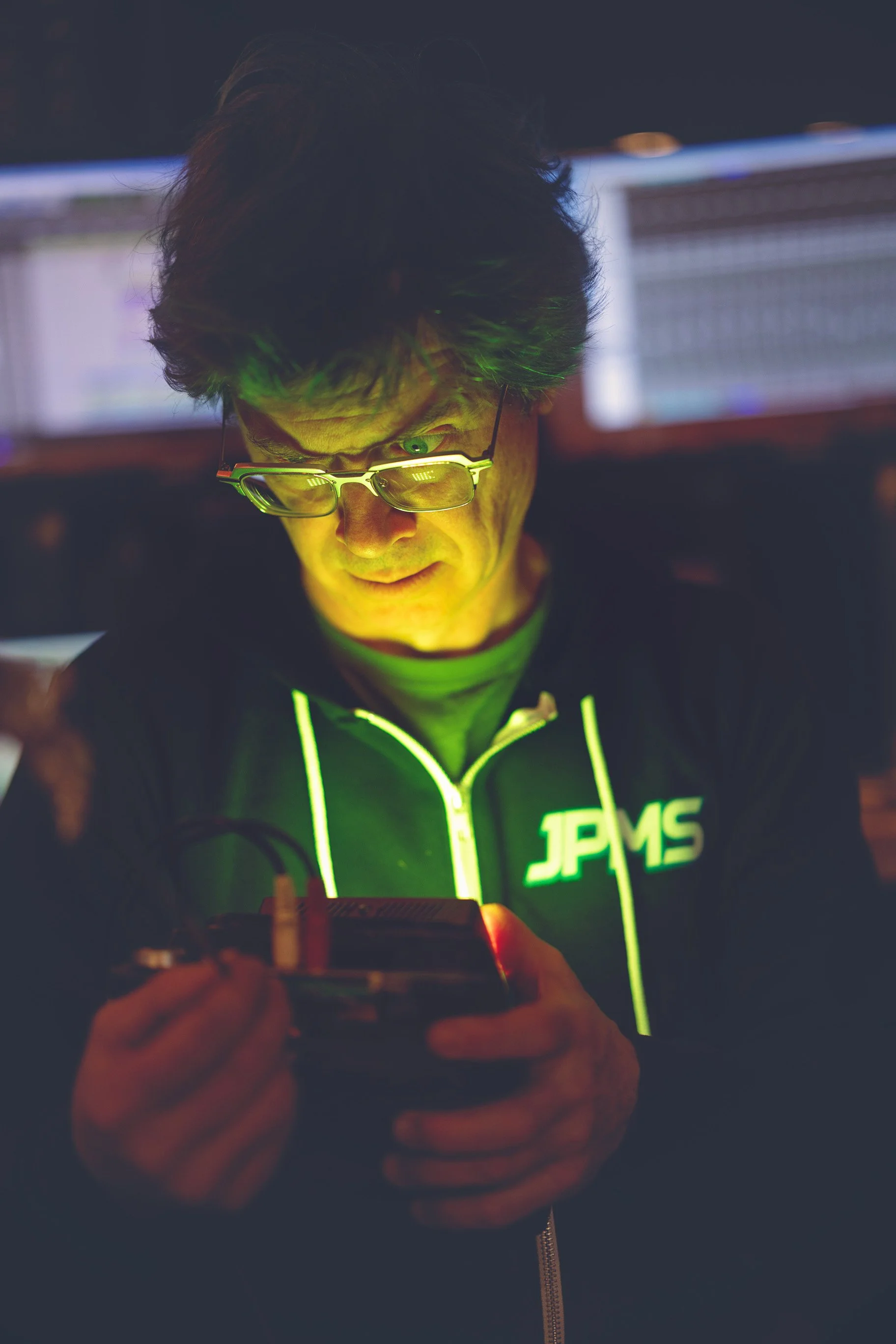 A man wearing glasses and a black jacket with neon green accents and the logo 'JPM6' on it, looking down at a device in his hands in a dimly lit environment with computer screens in the background.
