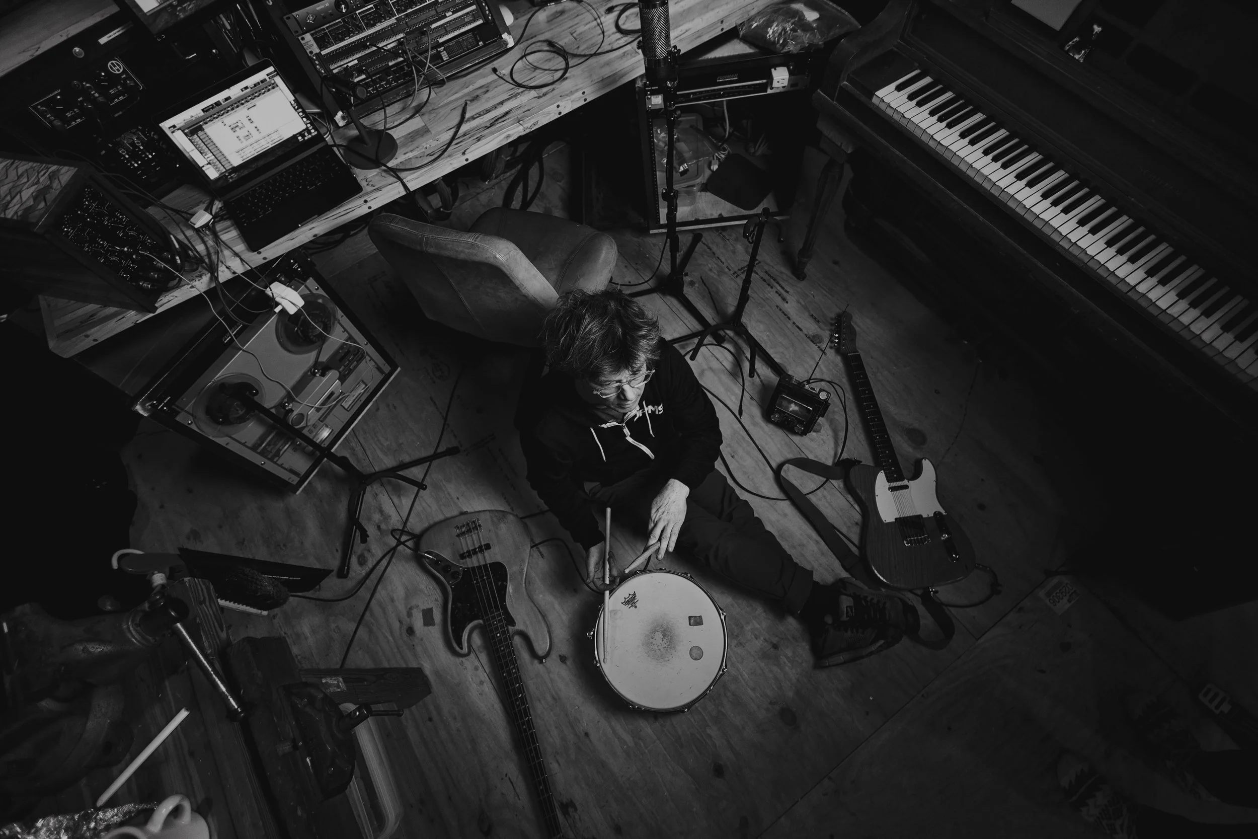 A person sitting on the wooden floor in a music studio, surrounded by musical instruments and electronic equipment, seen from above in black and white.
