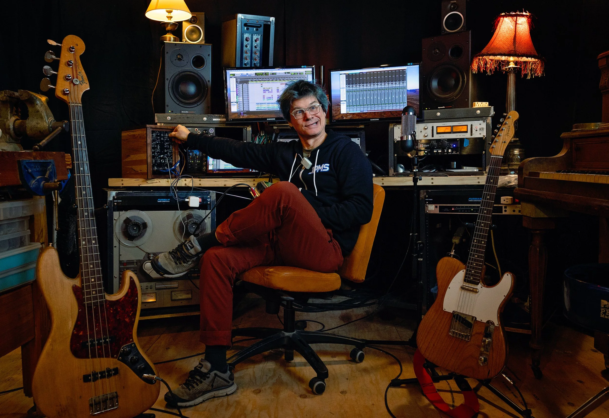 A man sitting in a music studio surrounded by musical instruments, including two electric guitars on either side, with audio equipment and computer monitors displaying audio editing software in the background.