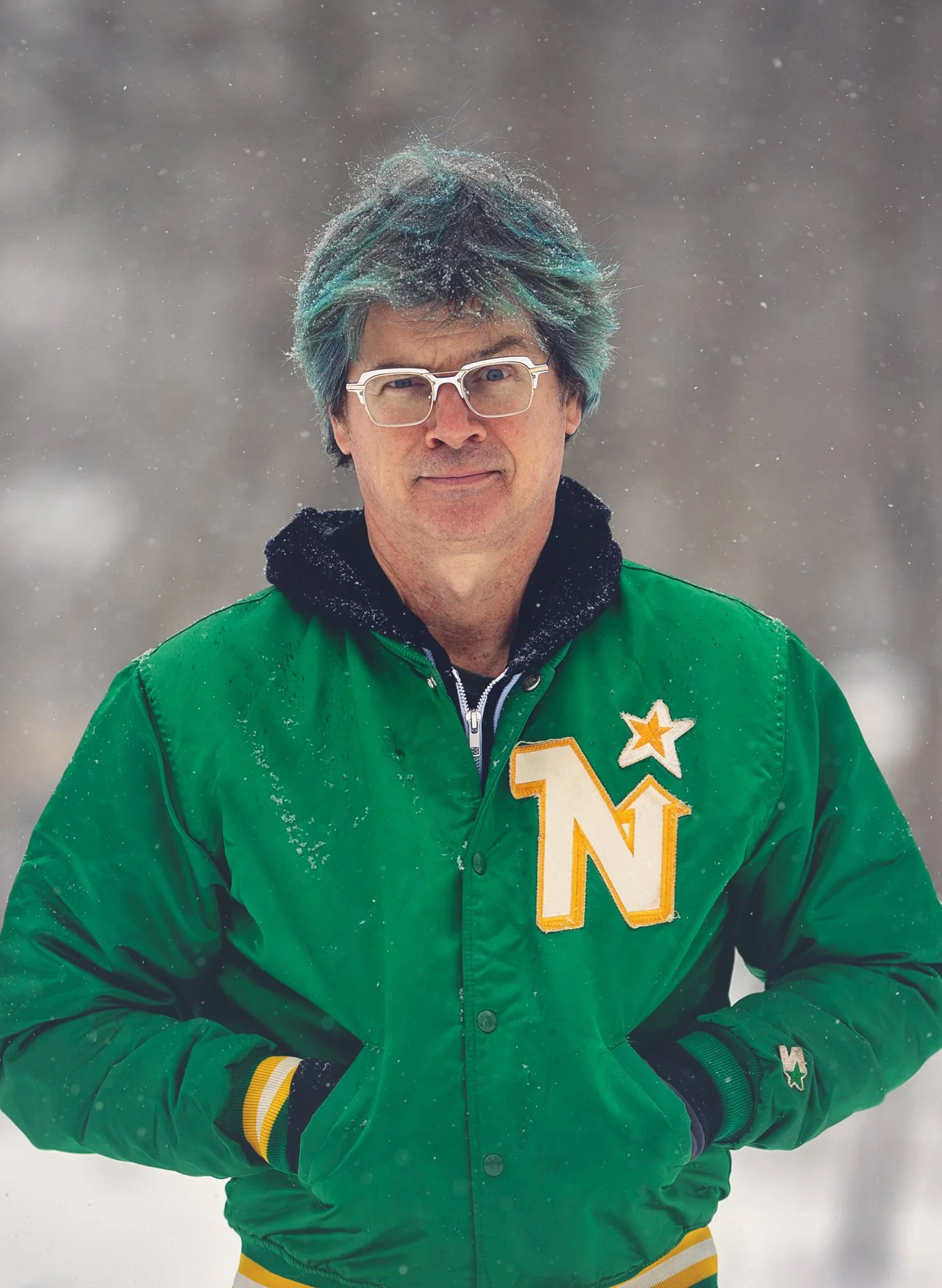 Man standing outdoors in snow, wearing glasses, a green varsity jacket with a large letter 'Northstars' and star patch, with his hands in pockets.