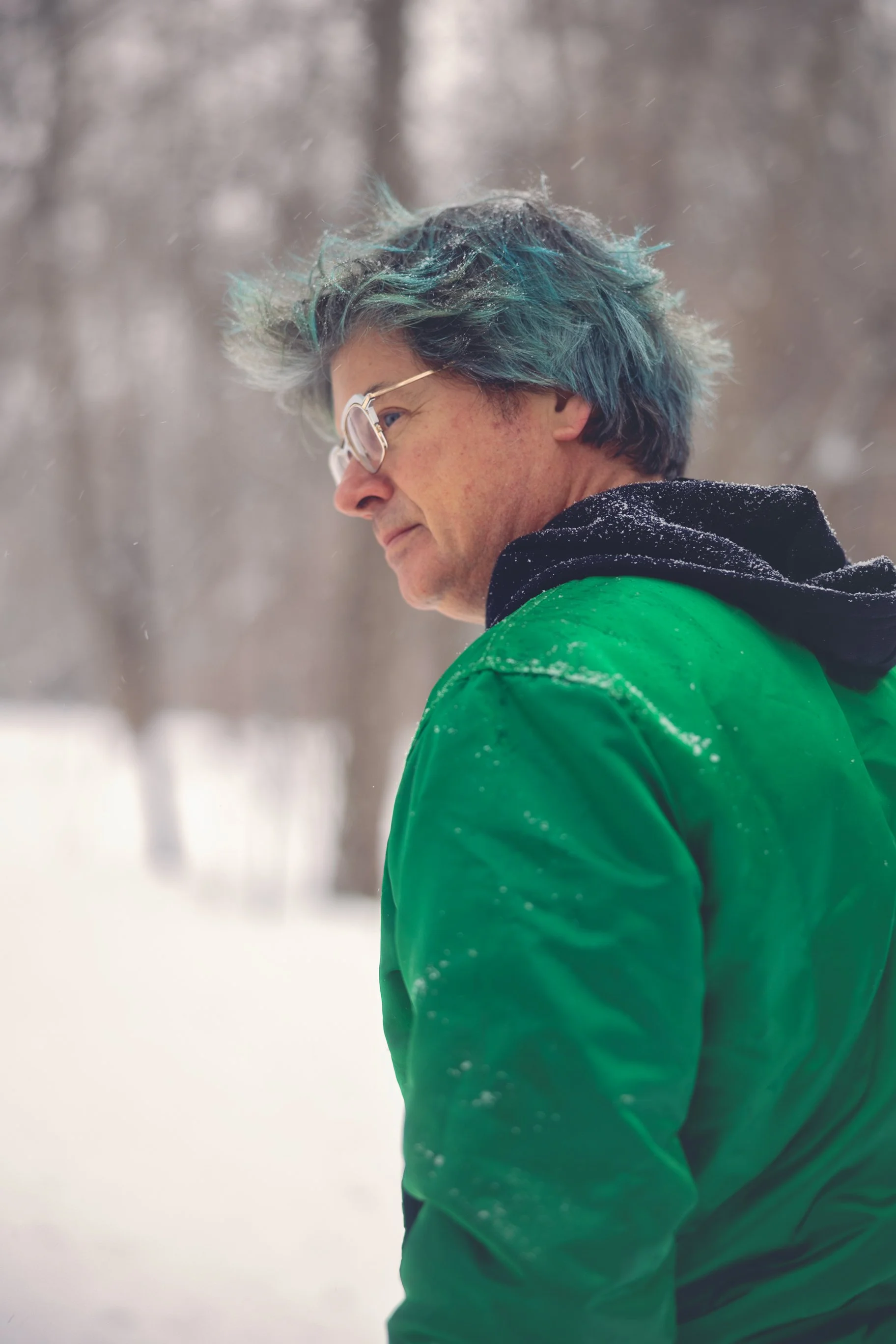 A person with blue-tinted hair wearing glasses and a green jacket, standing outdoors in a snowy environment, looking to the side.