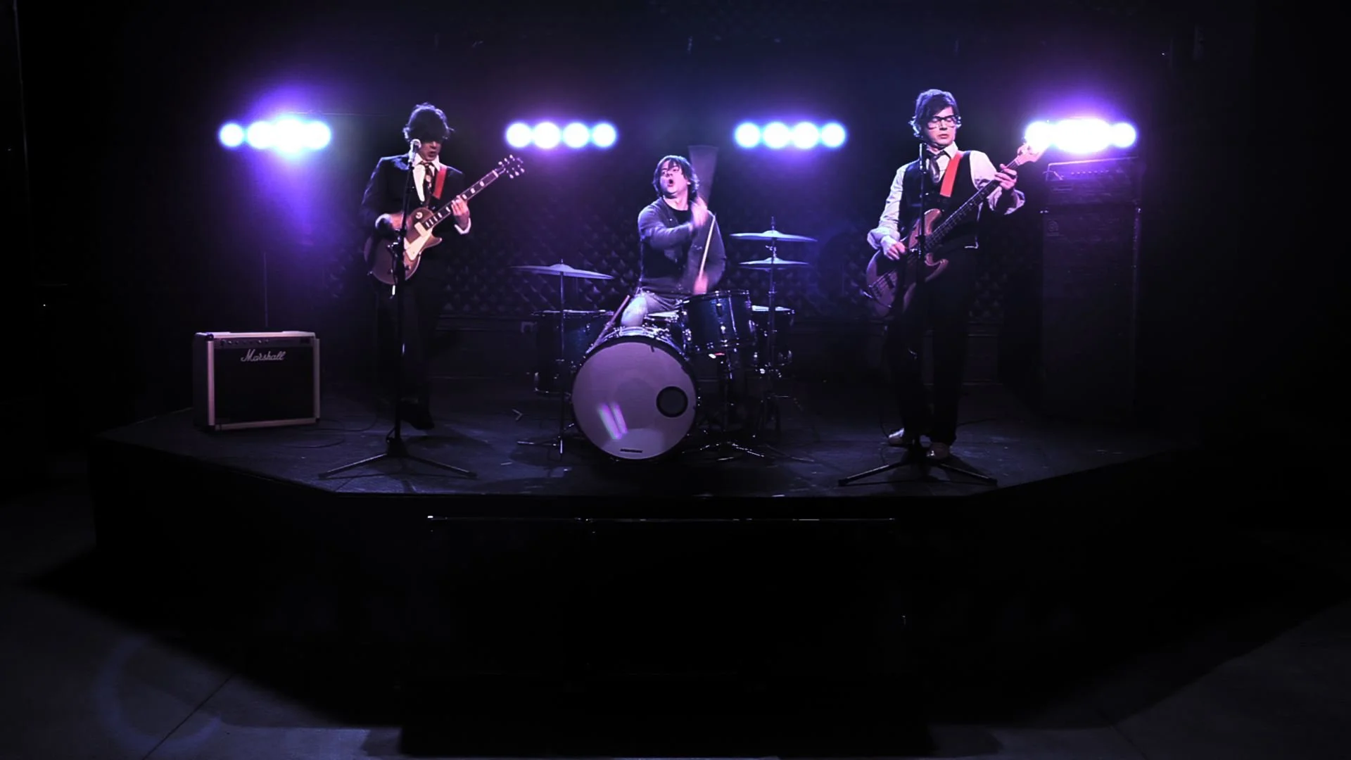 A band of three musicians performing on a dark stage with purple lighting. The band includes a guitarist on the left, a drummer in the center, and a bassist on the right. The guitarist and bassist are dressed in formal black suits with red accents, and the drummer is dressed casually in dark clothing.