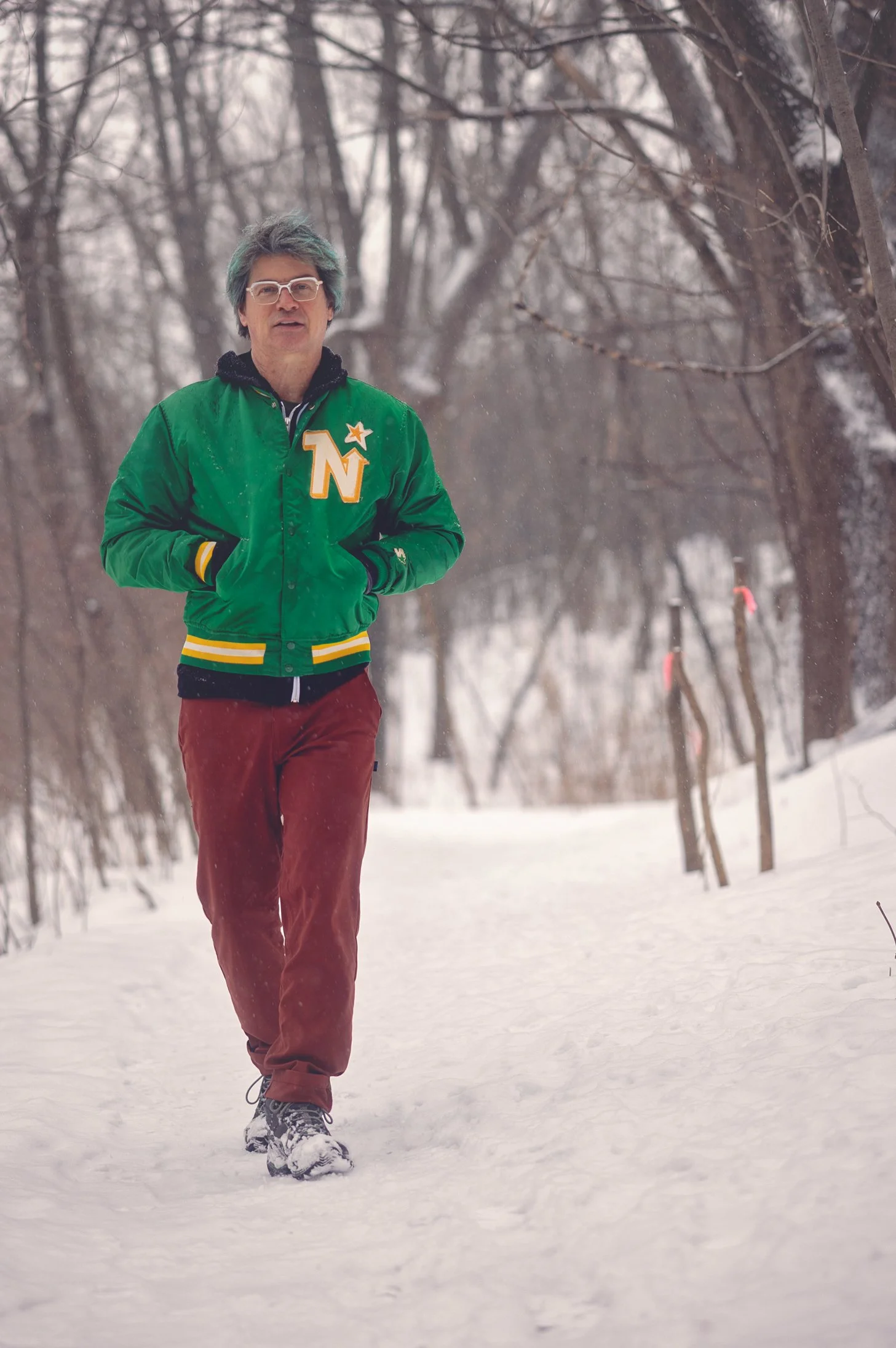 A person walking on a snow-covered trail in a winter forest, wearing a green 'Northstars' jacket, maroon pants, and snow boots.