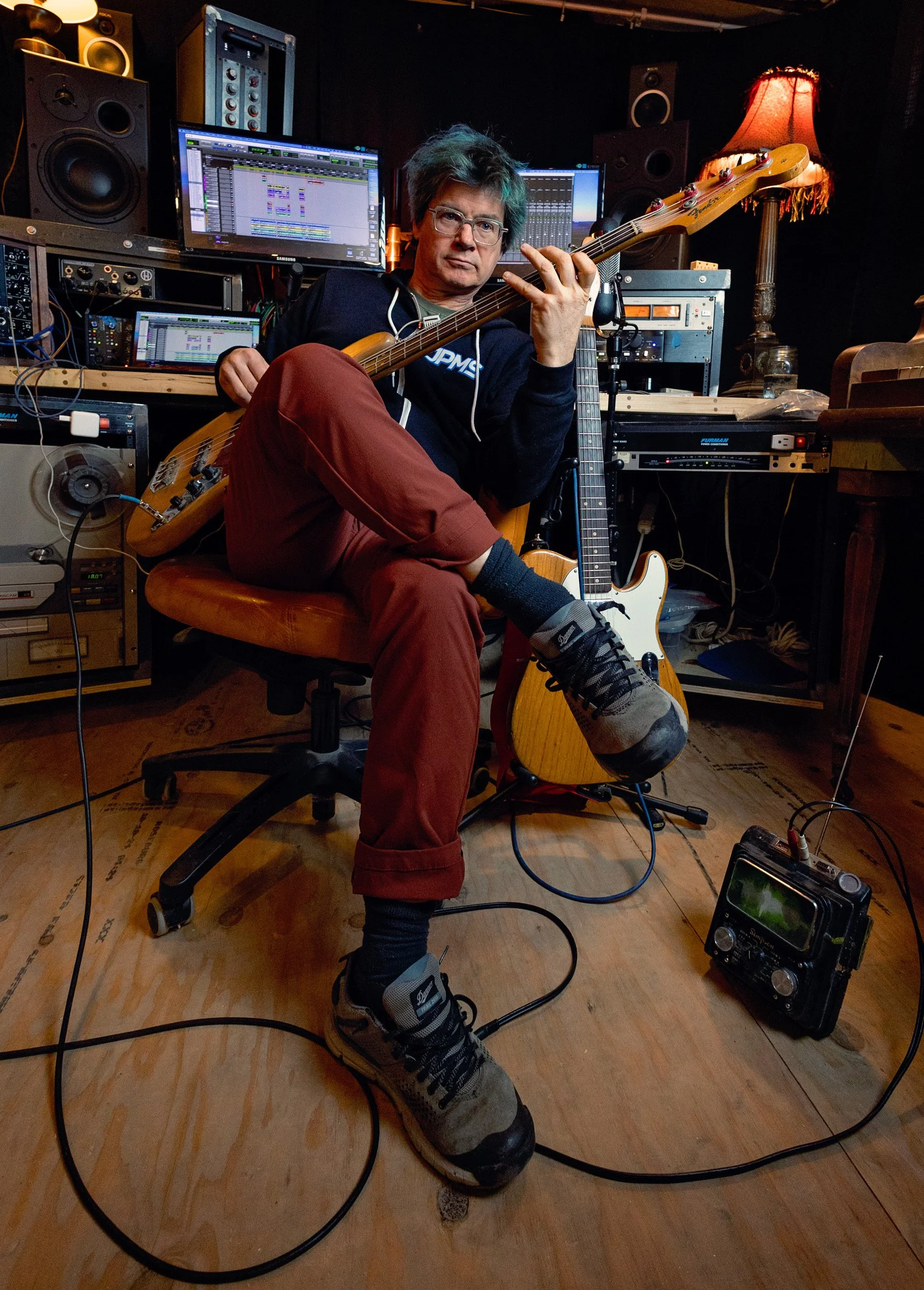A man with glasses playing an electric bass guitar in a recording studio surrounded by music equipment, computers, and speakers.