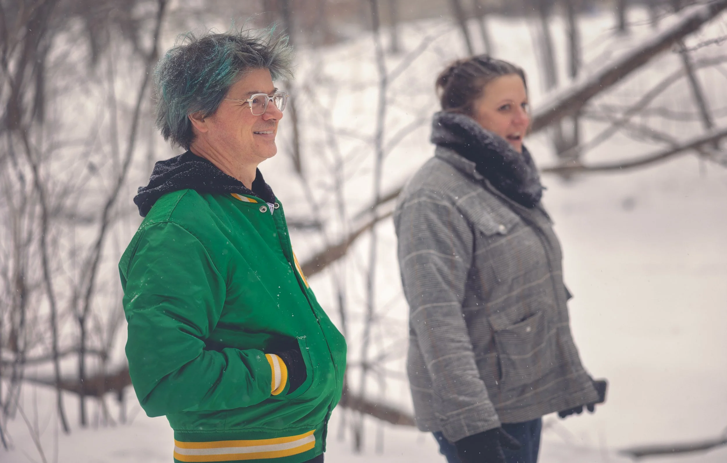 Two people standing outdoors in a snowy landscape, wearing winter clothing. The man on the left has glasses and multicolored hair, wearing a green jacket. The woman on the right has her hair in a bun and is wearing a gray jacket and black gloves.