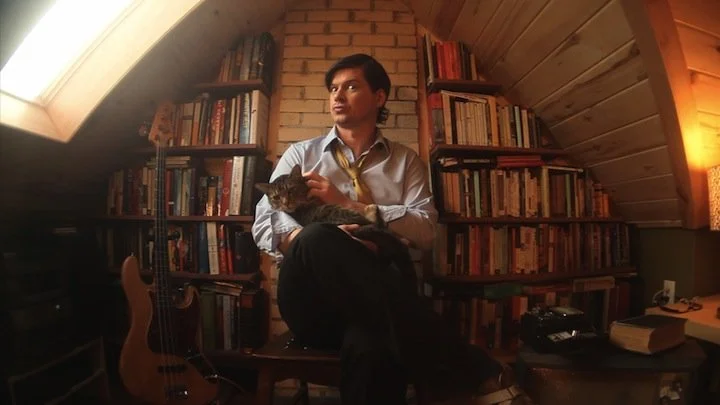 A man sitting on a wooden bench in a cozy room with sloped ceiling and brick wall, holding a cat while bookshelves filled with books are in the background.
