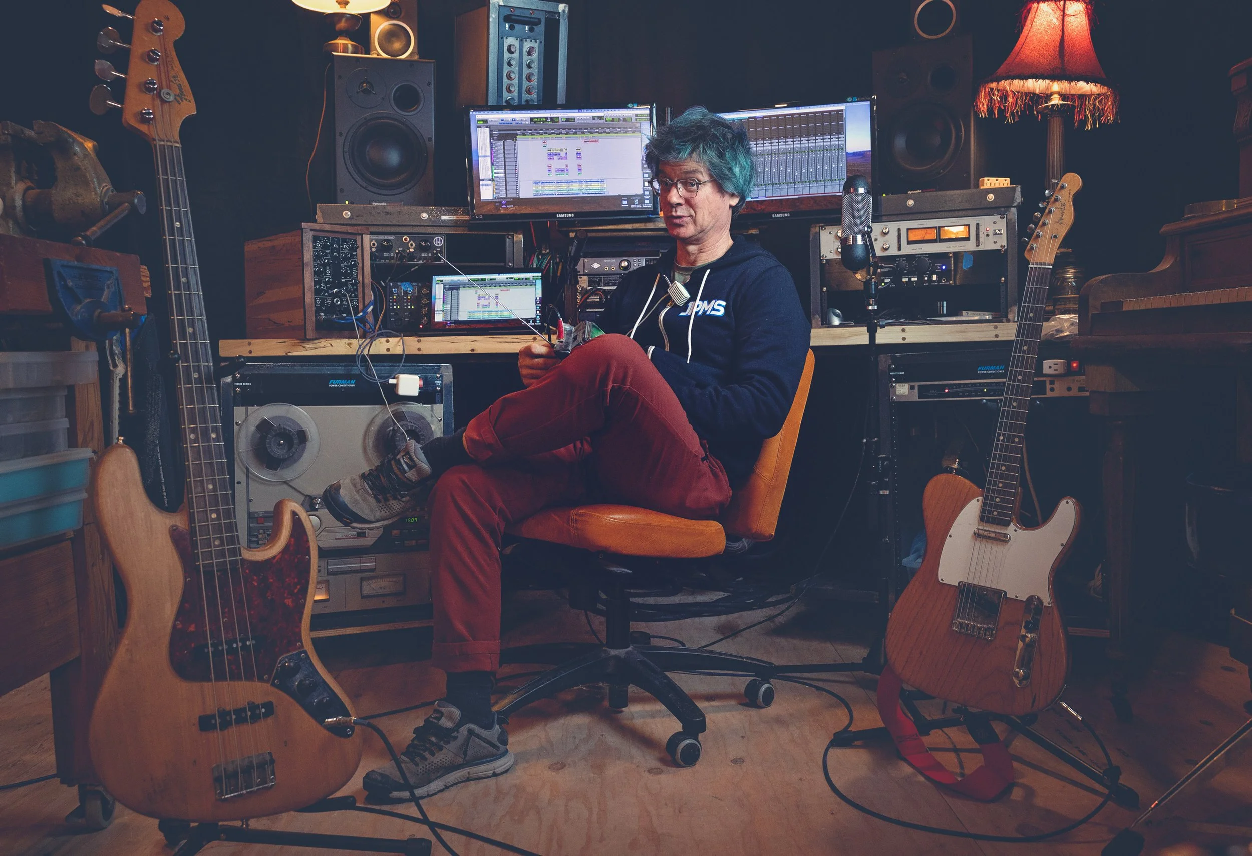 A man sitting in a music studio surrounded by guitars, computers, and audio equipment, working on music production.