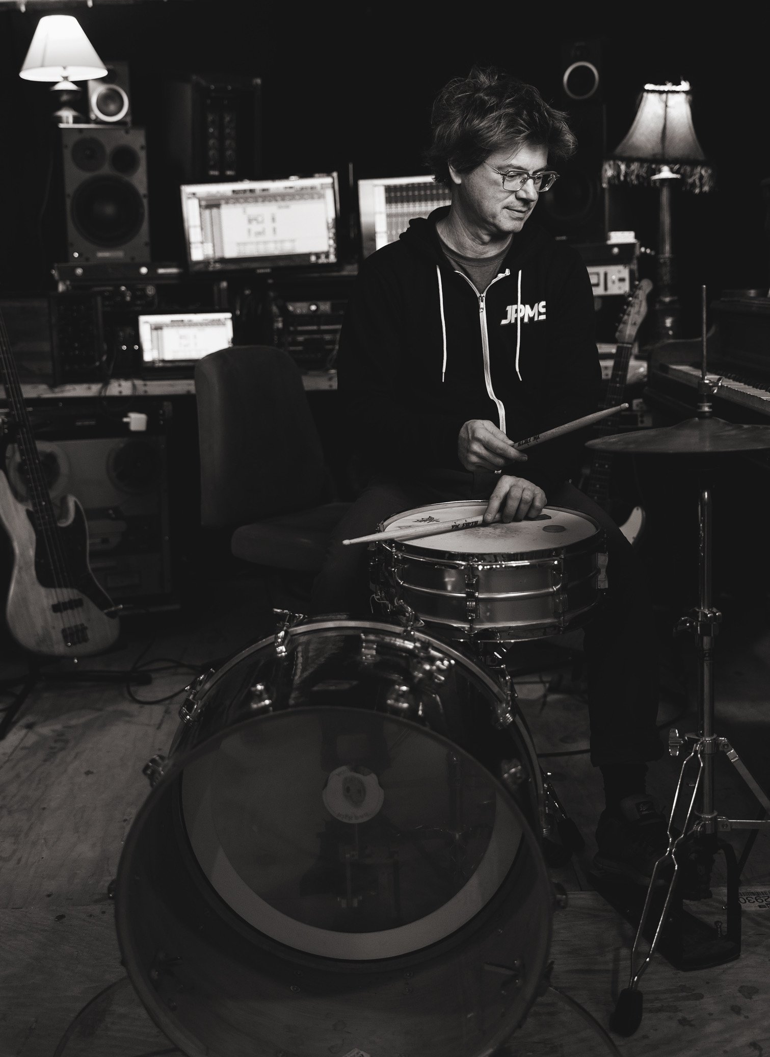 A man sitting in a recording studio playing a drum set with electronic equipment and guitars in the background.