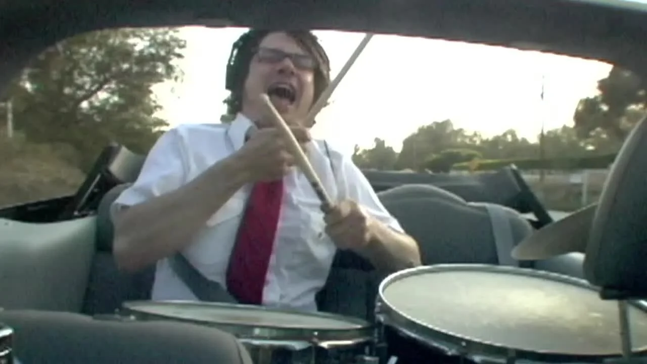 A man with glasses and messy hair, wearing a white shirt with a red tie, sitting in a car, holding a drumstick, and singing passionately with an open mouth while playing drums.