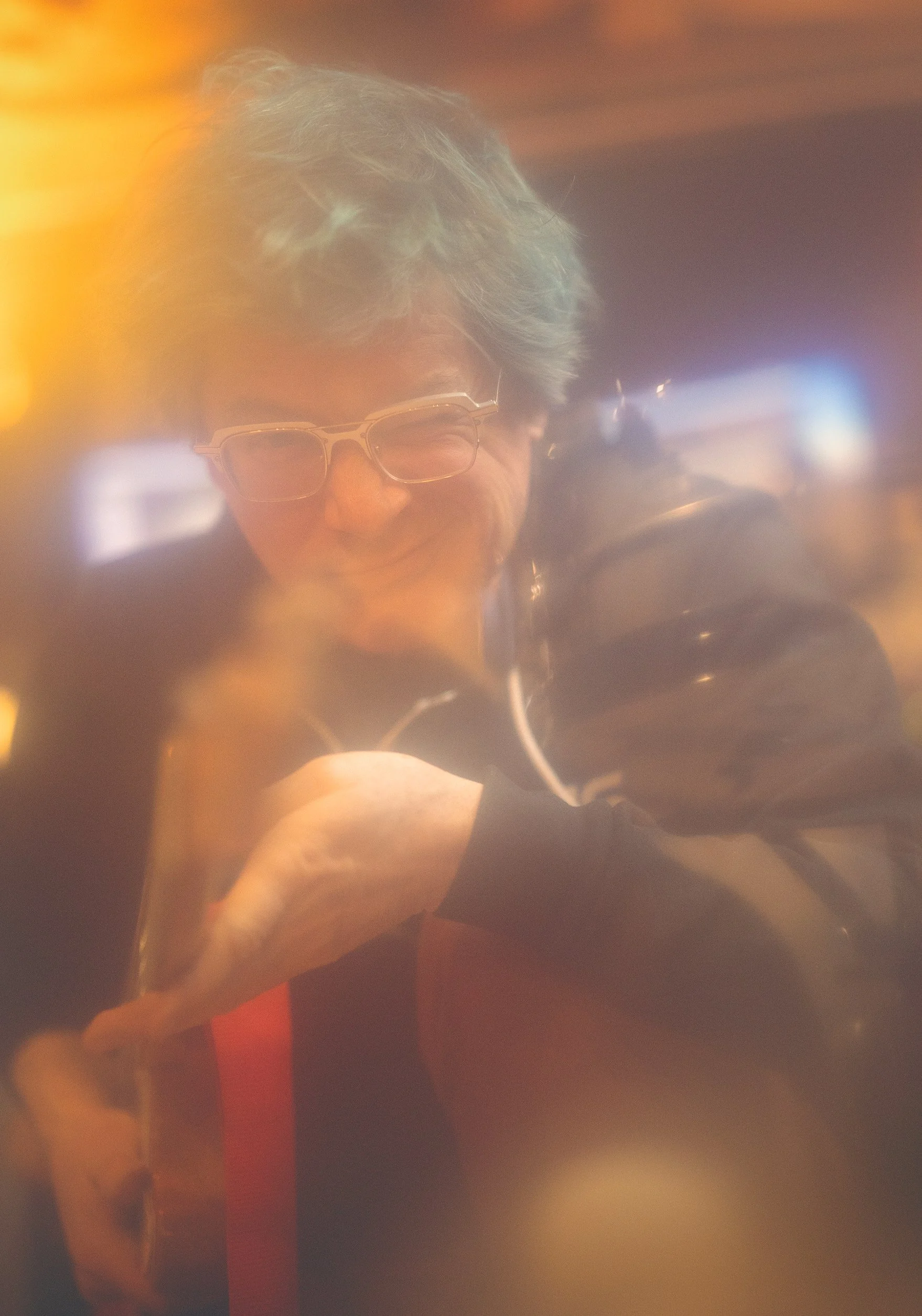 A person with gray hair and glasses smiling, holding a beverage, with a soft-focus, warm-toned background.