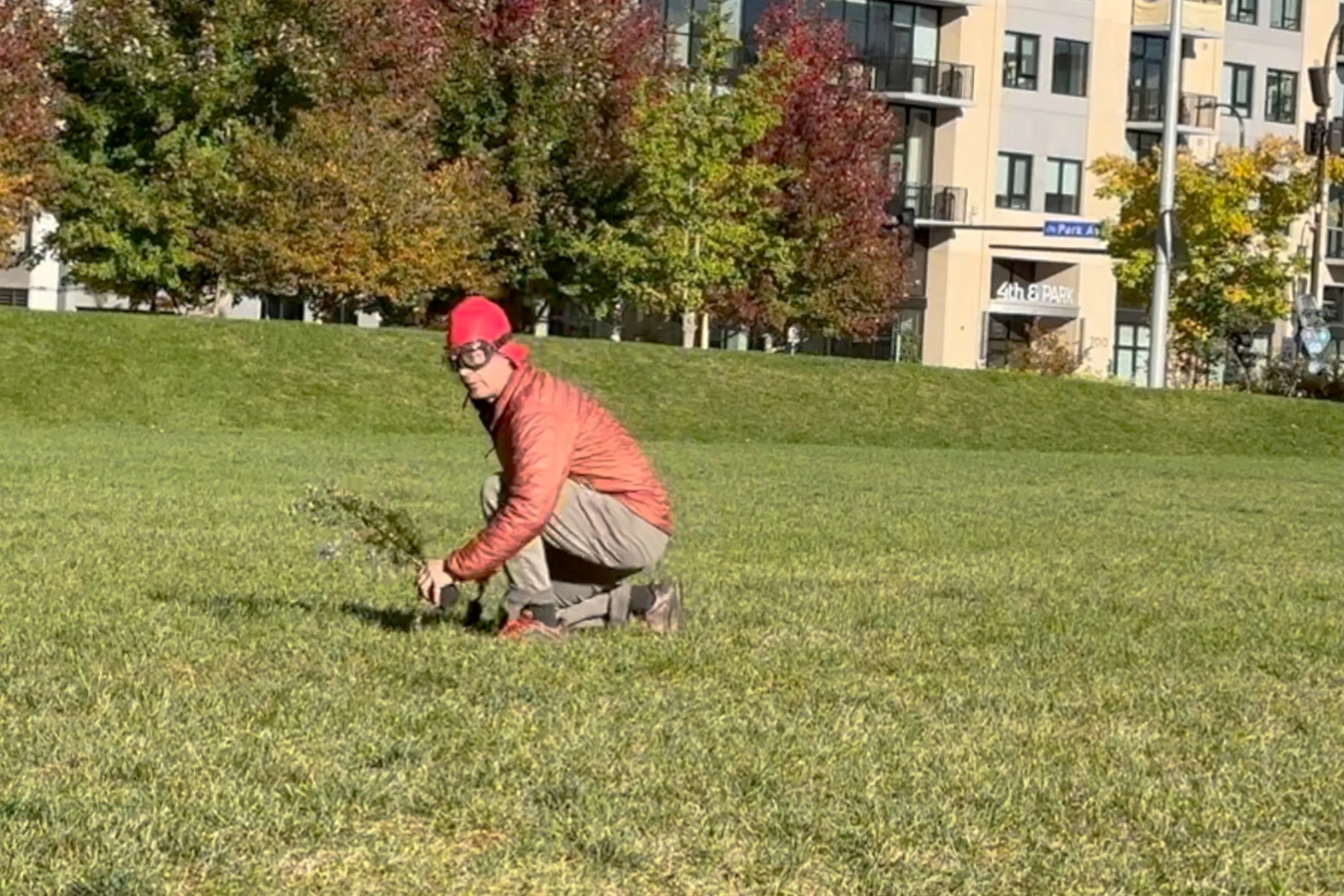 A person positioned close to the ground on a grassy field, wearing a red jacket, red cap, glasses, beige pants, and brown shoes, with trees and a modern apartment building in the background.