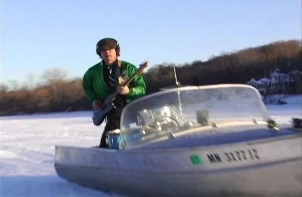 A person wearing a green jacket, black headphones, and glasses playing an electric guitar next to a boat on a snowy landscape.