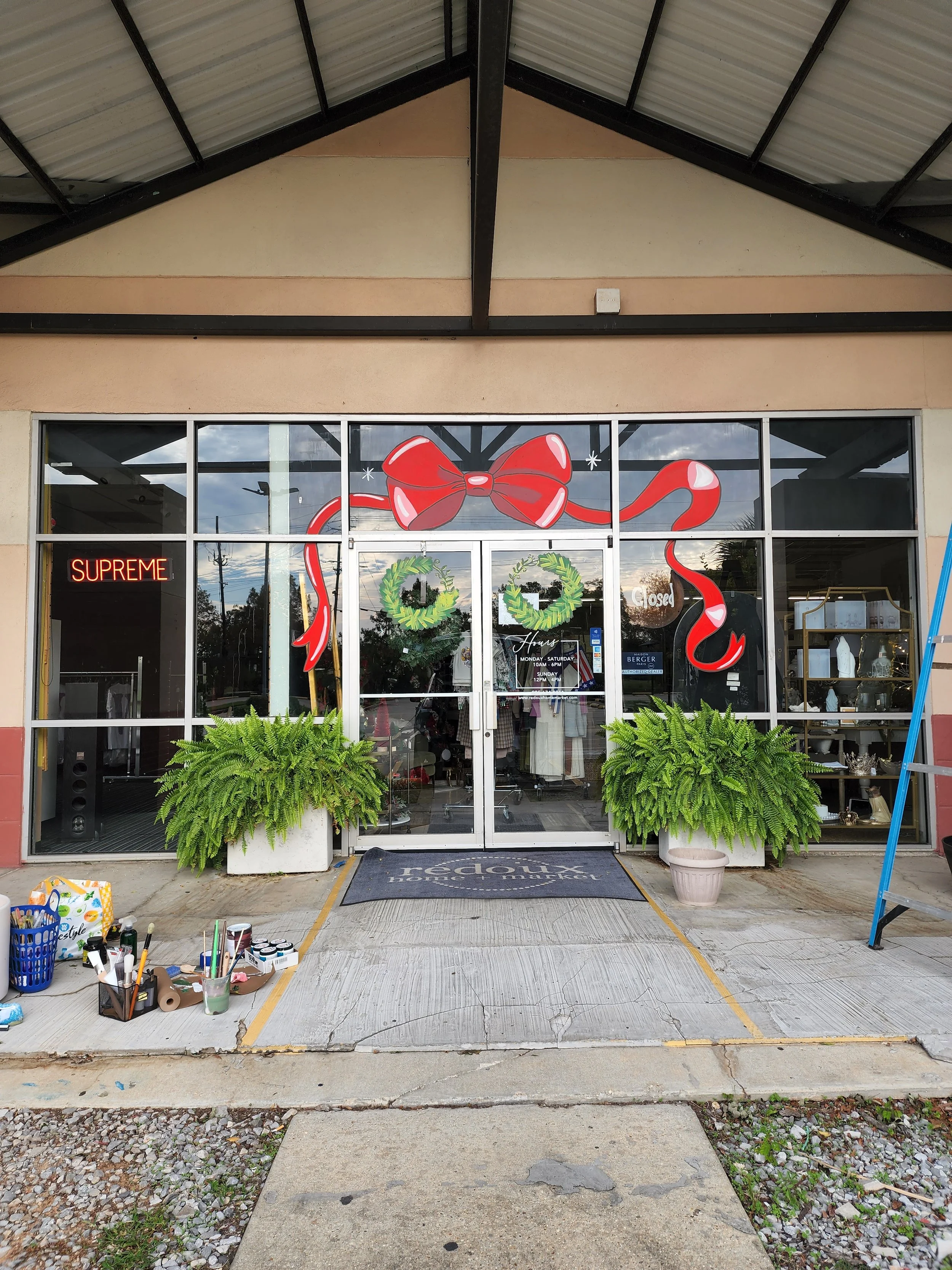 Storefront decorated with a large red bow and ribbon, two green wreaths hanging on the door, potted ferns outside, and an indoor display visible through glass doors. There are also various items on the sidewalk outside.