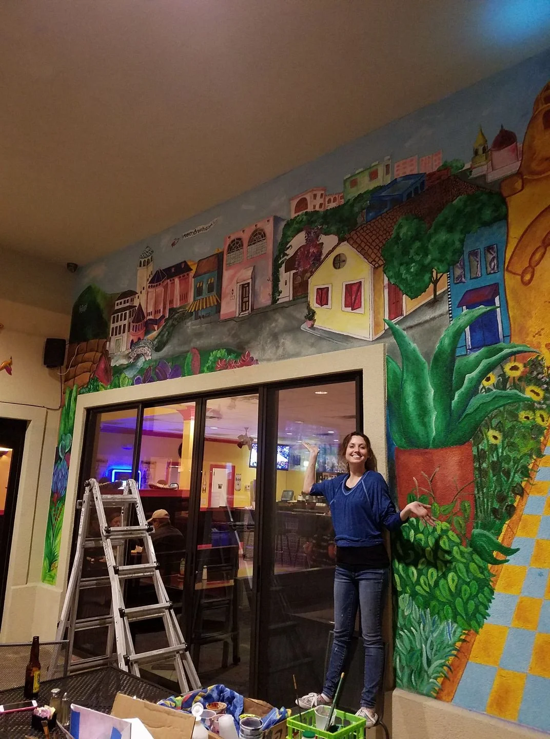 Woman smiling and posing enthusiastically next to a colorful mural of buildings and plants on the wall of a restaurant or cafe.