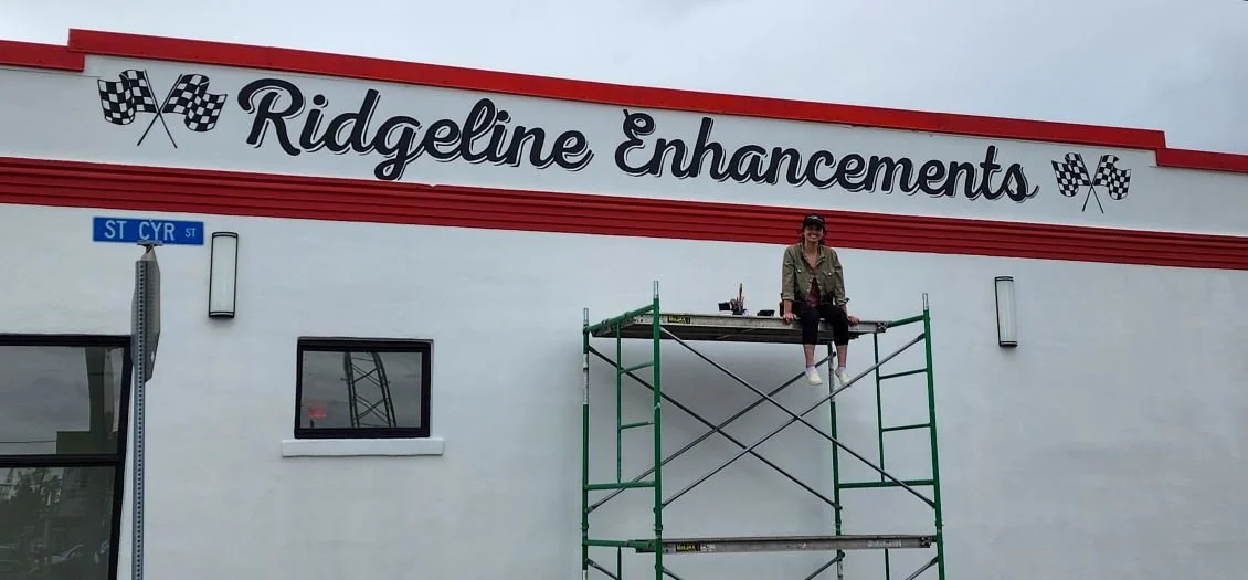 Person sitting on scaffolding in front of a building with a sign that reads "Ridgeline Enhancements" and checkered flags. The building has a white facade with red trim, and a street sign indicating "ST CYR ST."