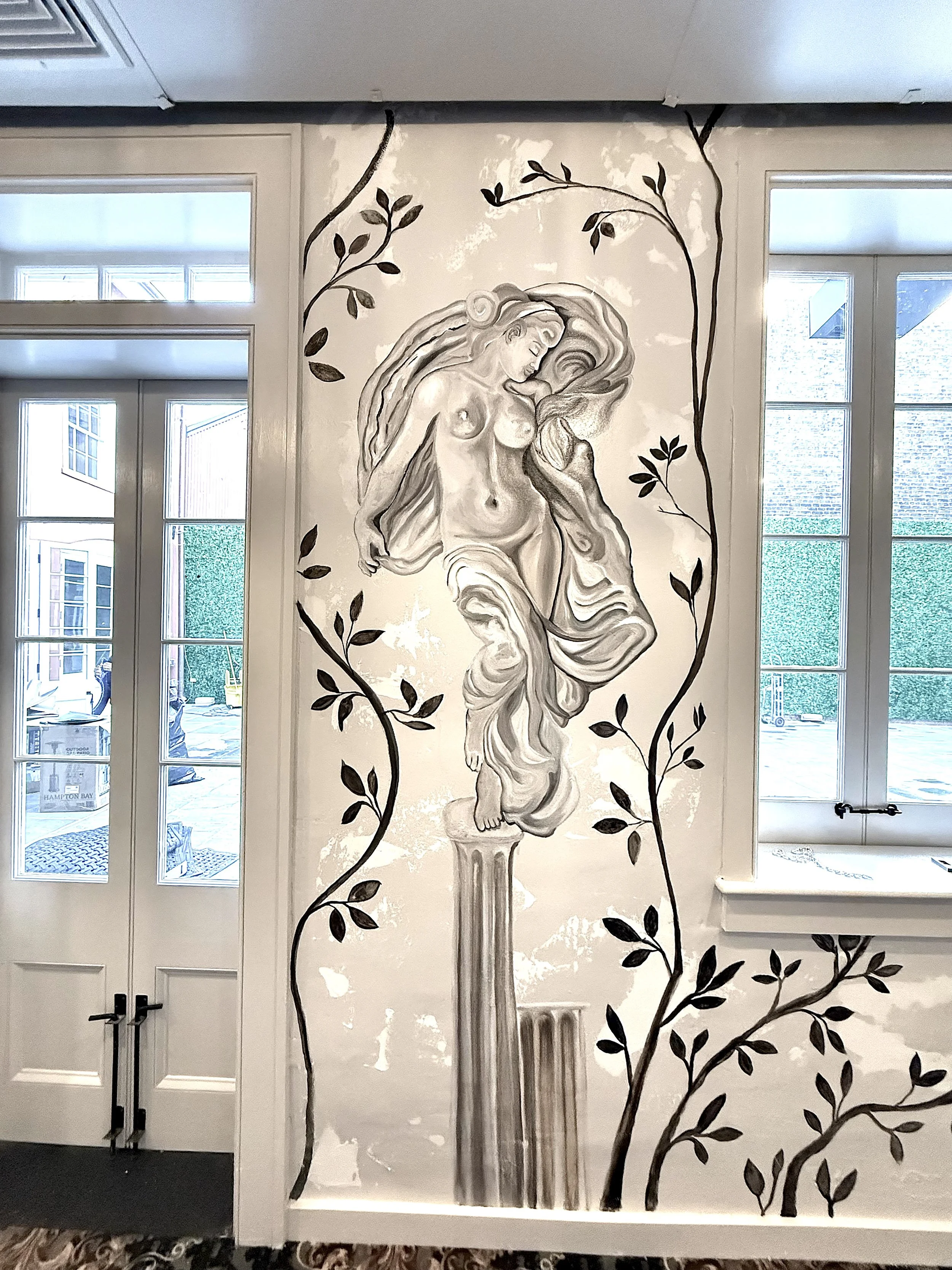 A monochrome mural of a classical sculpture of a woman on a pedestal, surrounded by black painted tree branches and leaves, on an interior wall near a glass door and window.