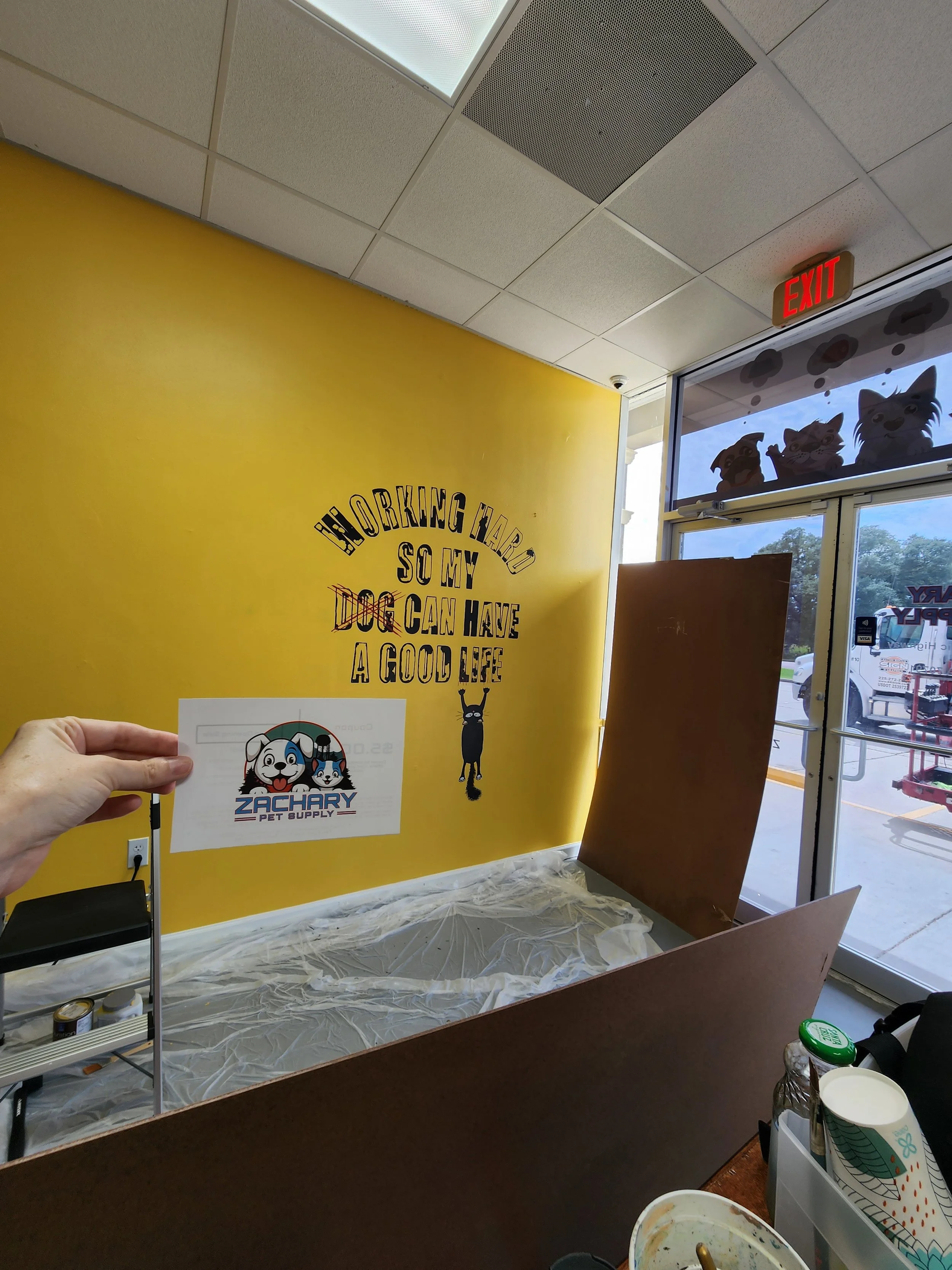 An interior view of a pet store with a yellow wall, a sign that reads "Working hard so my dog can have a good life," and a window with cartoon cat silhouettes. A hand holds a Zachary Pet Supply flyer in the foreground.