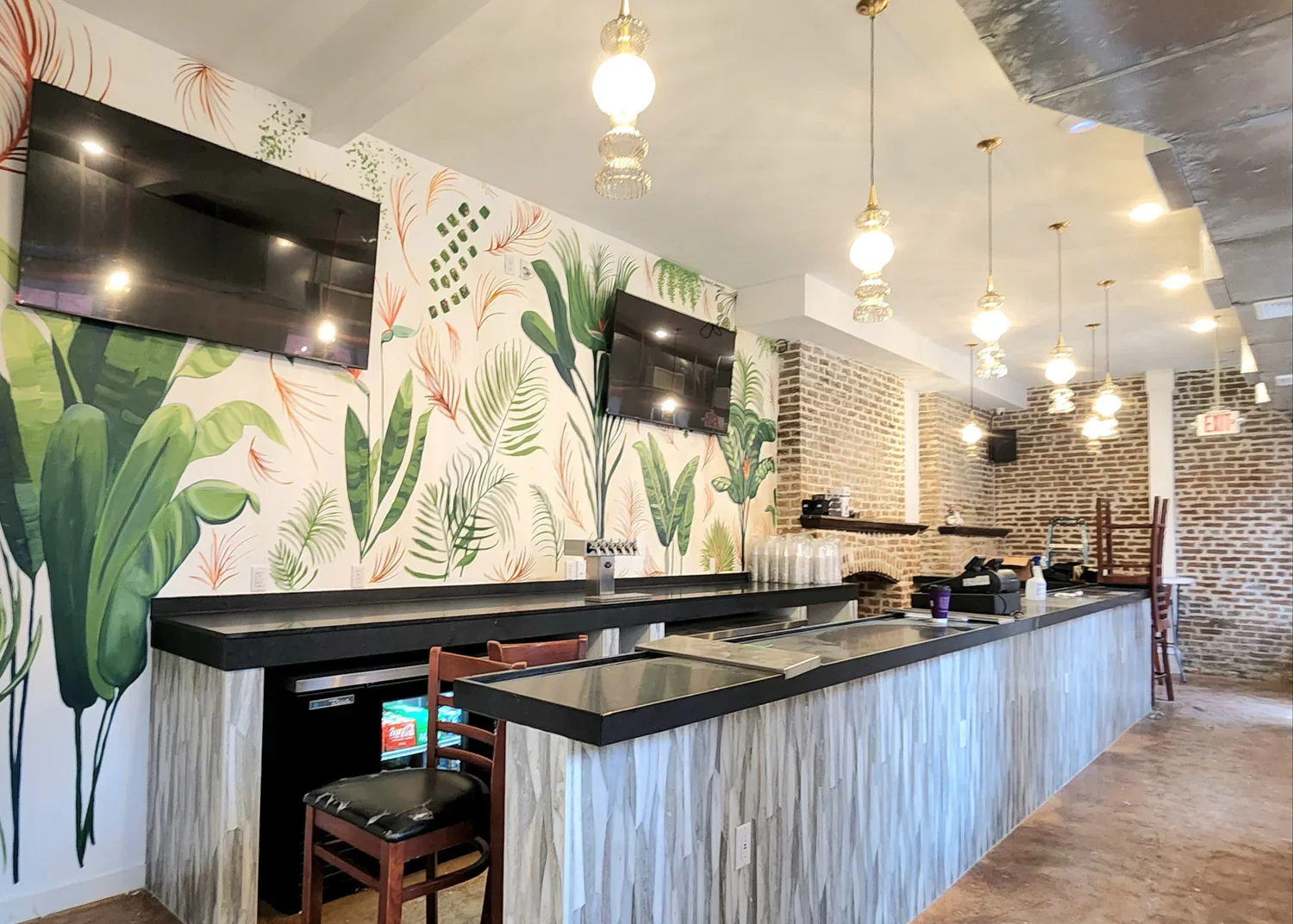 Interior view of a restaurant or cafe with a tropical-themed wall mural, black countertop bar, and pendant lights hanging from the ceiling. There are two flat-screen TVs on the wall and items on the bar including glasses, a cash register, and beverag