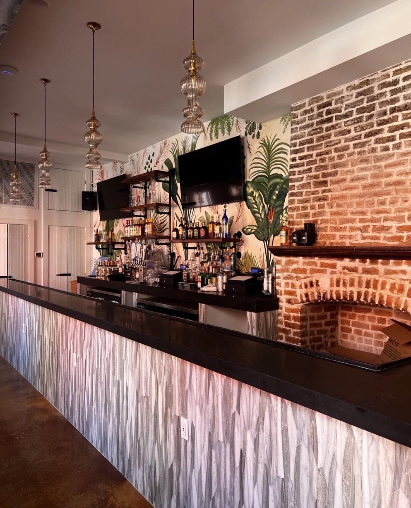 Indoor bar with tropical-themed wallpaper, a black marble counter, and a brick wall with a fireplace. Shelves above the bar hold liquor bottles, and two TV screens are mounted on the wall. Decorative pendant lights hang from the ceiling.