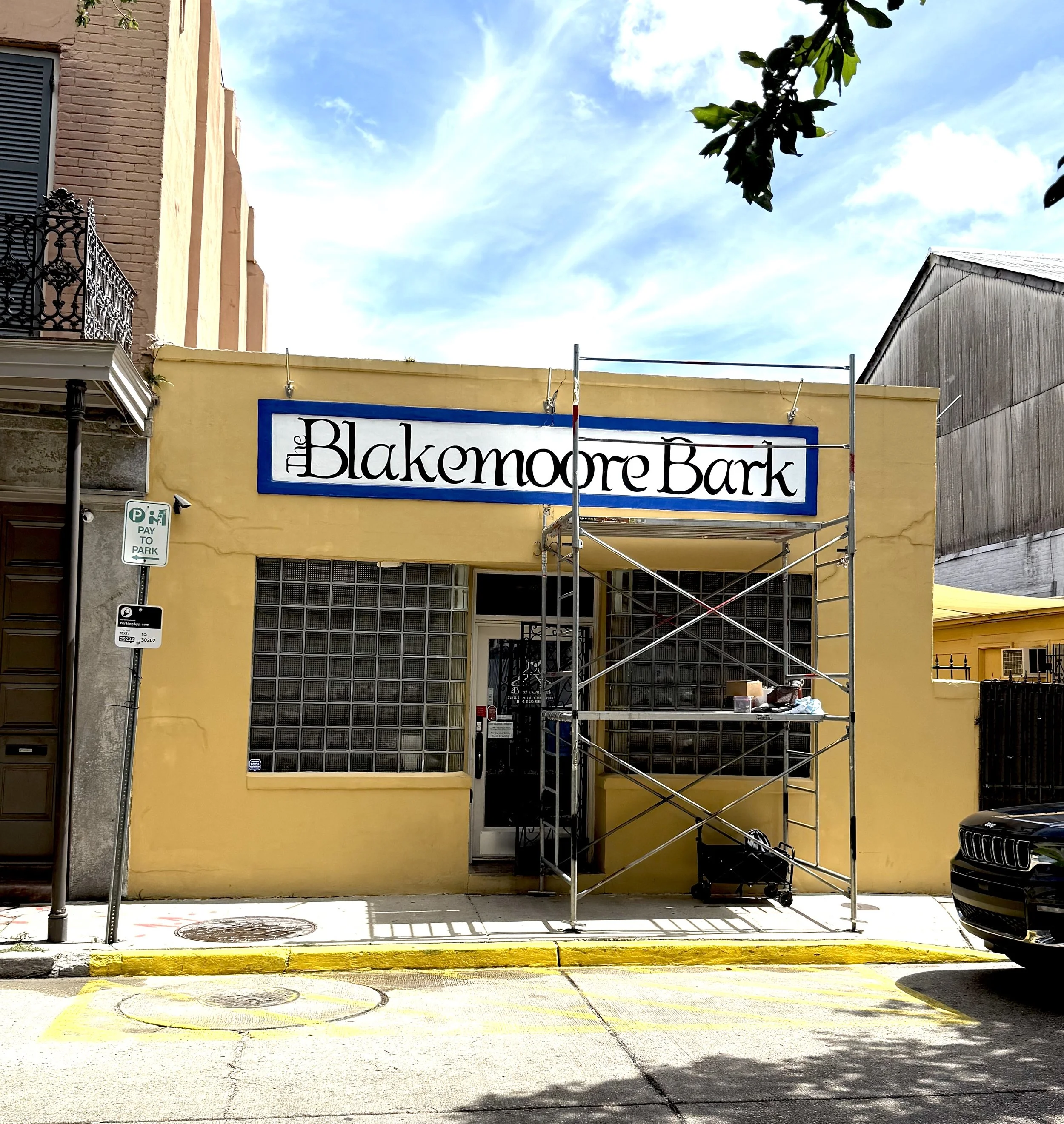 Building with a yellow exterior and a large sign that reads "The Blakemoor Bark" in black letters on a white background with a blue border, with scaffolding in front of the entrance, parked car on the street, and a parking sign on the left.