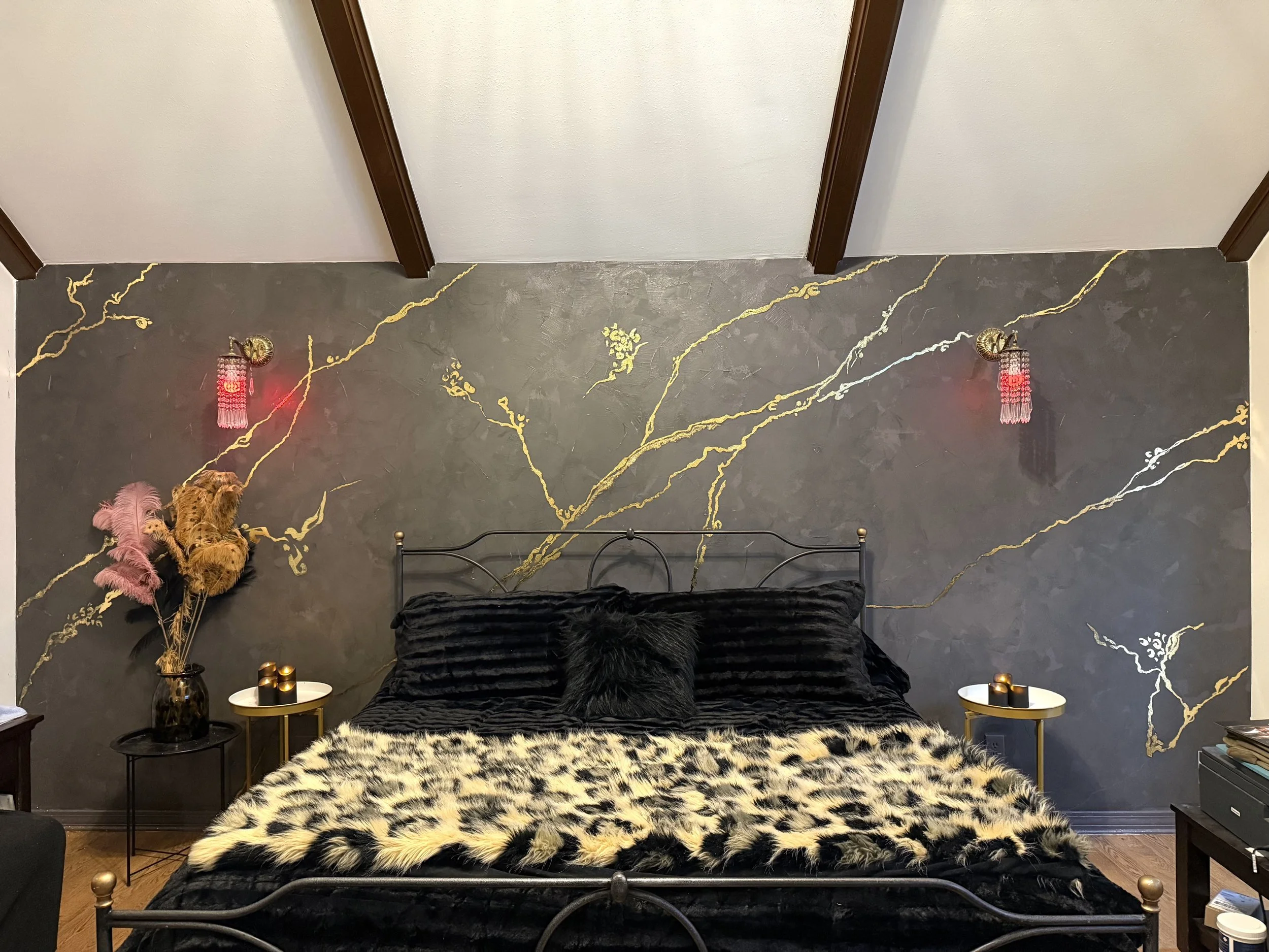 A bedroom with a black metal bed frame, black bedding, a furry black pillow, and a faux tiger fur blanket. The wall behind the bed has a dark marble pattern with gold veins, two pink wall sconces with tassels on either side, and a pink feather bouque