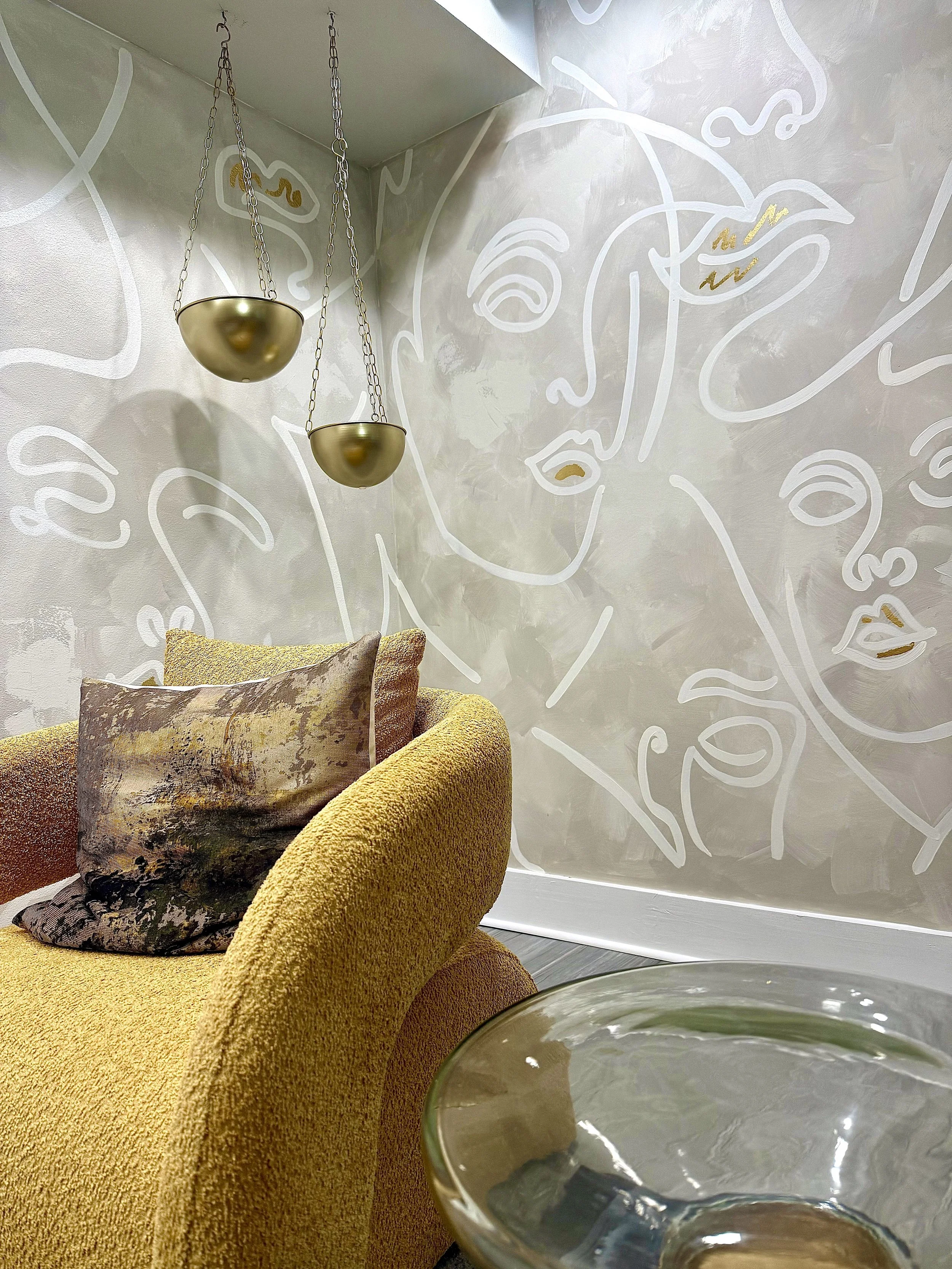 Interior of a modern lounge with a yellow armchair, decorative pillows, a glass side table, and a wall mural of abstract line art faces. Two hanging gold-colored bowls are suspended from the ceiling.