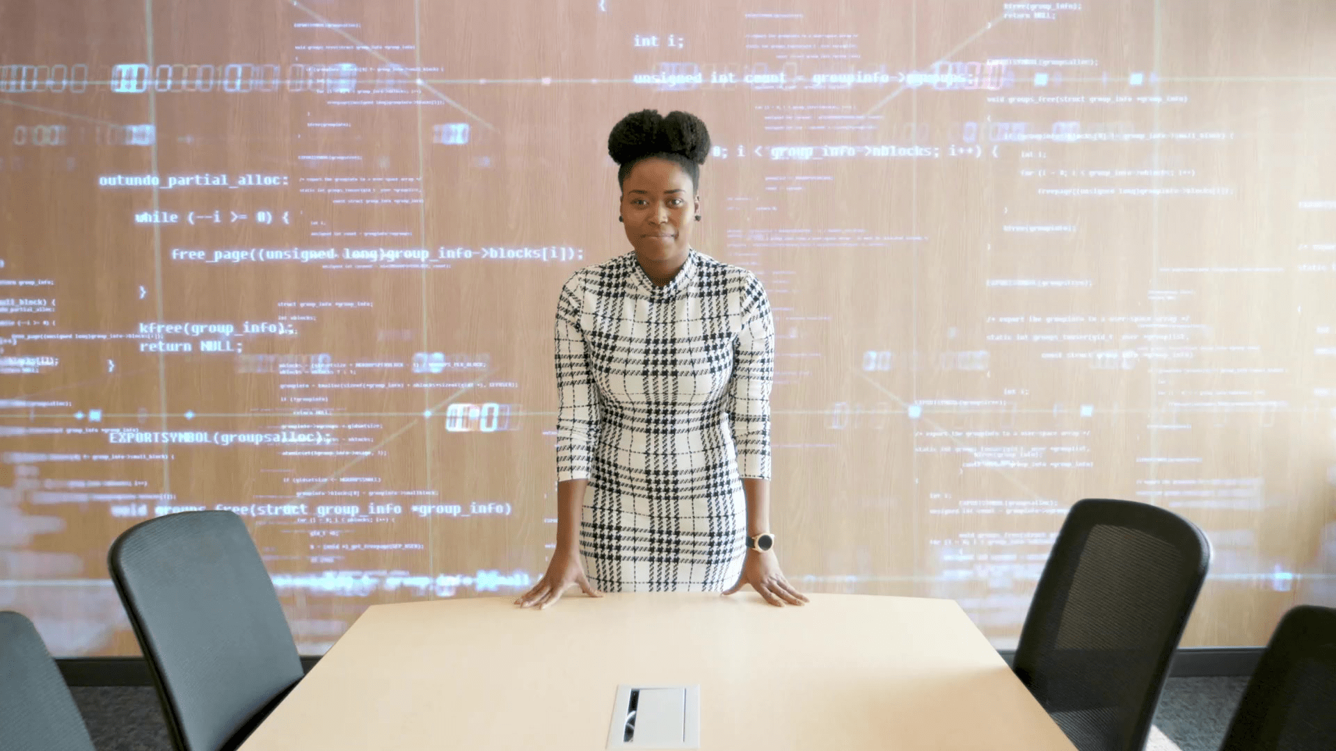 Woman standing behind a conference table, with a digital code projection on the wall behind her.