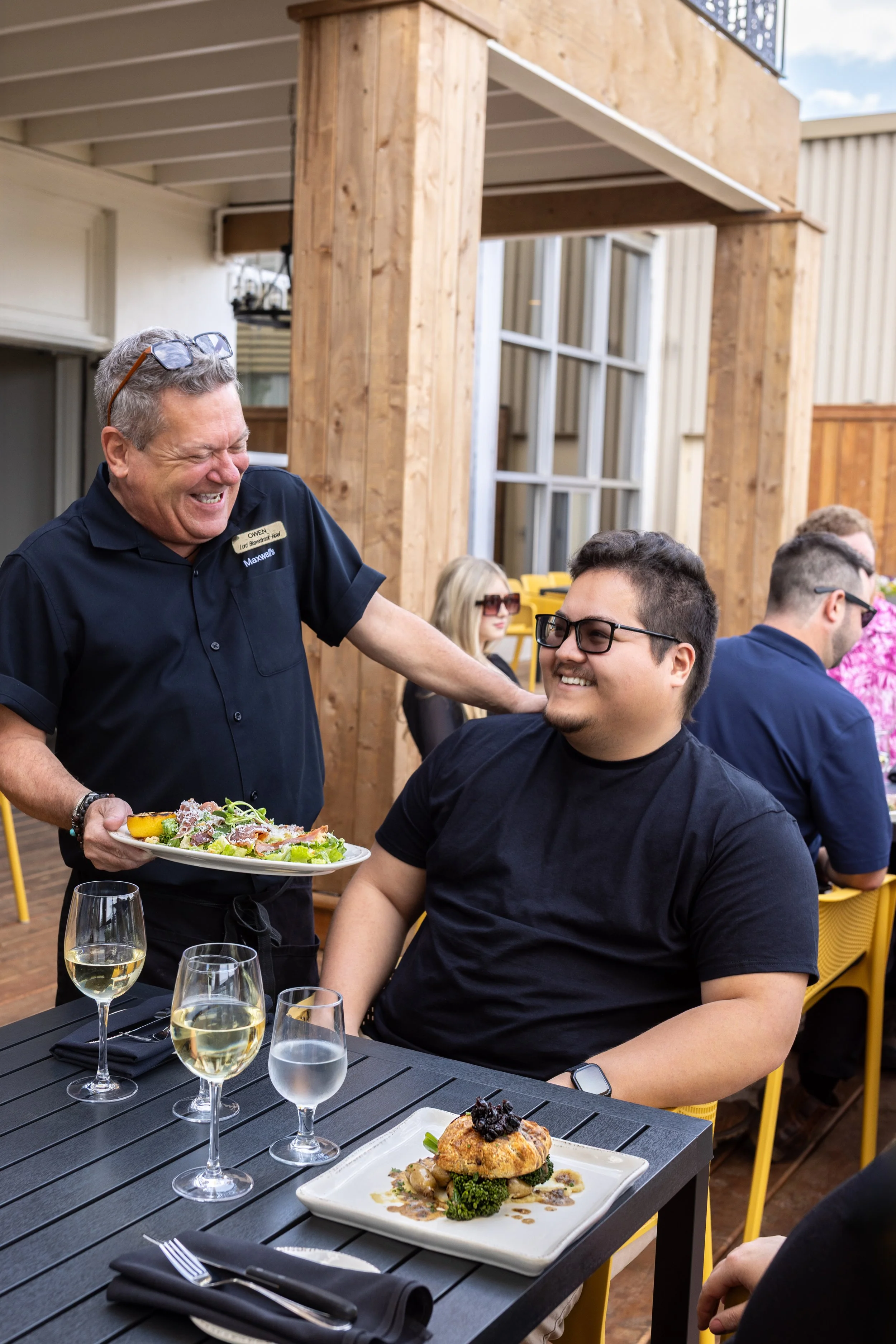 A man in a black shirt is seated at a table on Maxwell's Steak & Seafood patio with a plate of food, smiling at a server who is serving a salad. The server is laughing, wearing a black uniform.