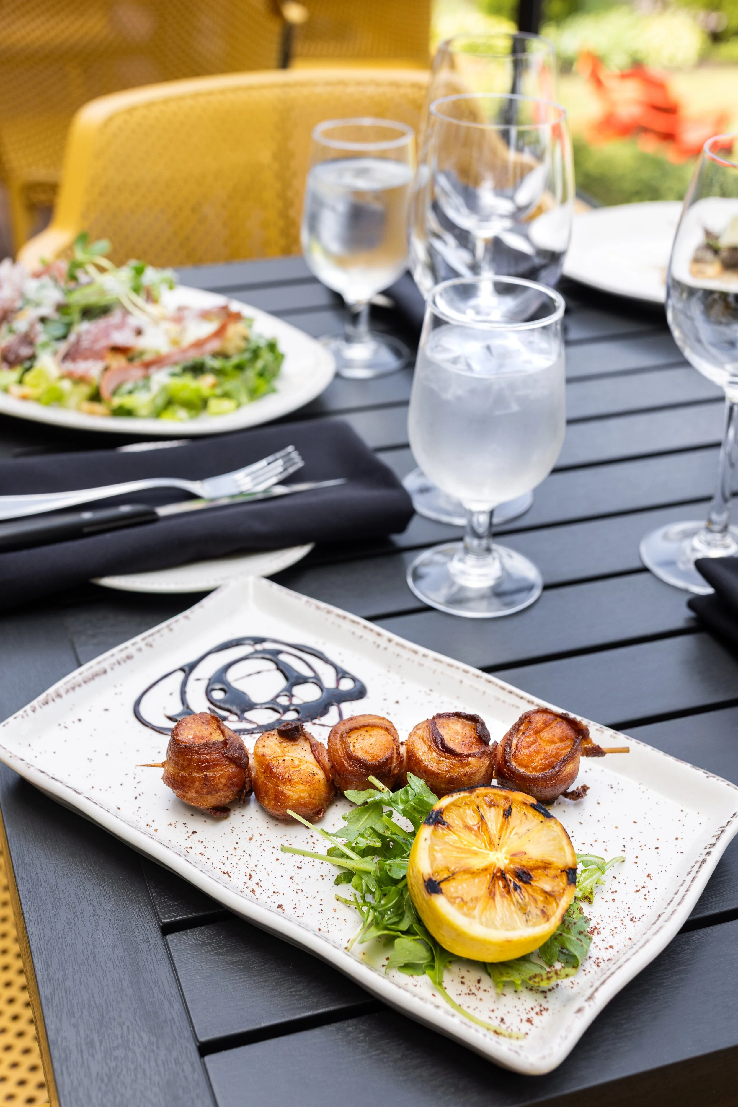 Plate of bacon-wrapped scallops grilled with lemon and balsamic drizzle on a black outdoor dining table of Maxwell's Steak & Seafood patio.
