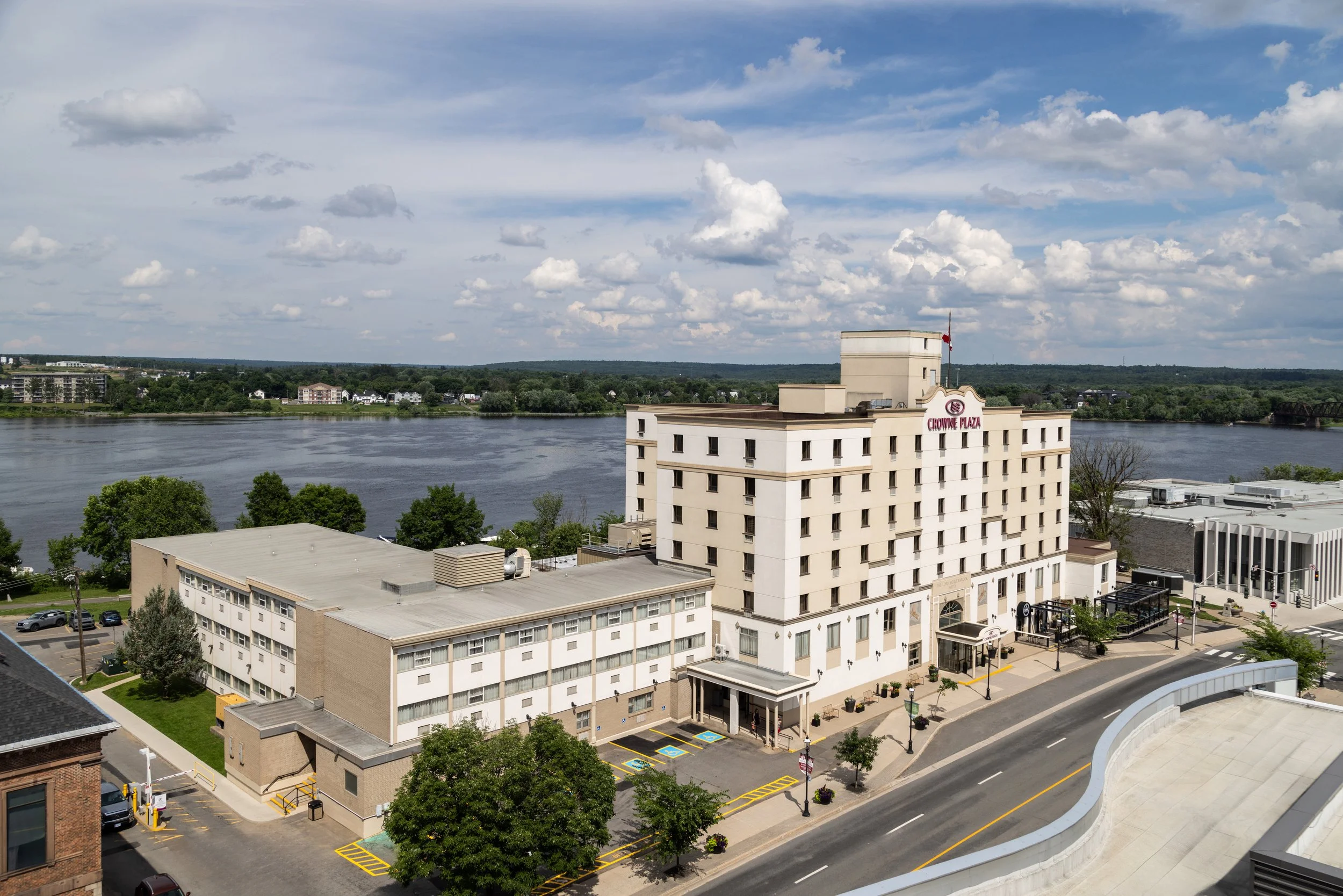 Aerial view of a hotel called Crowne Plaza Fredericton situated near the Saint John river with trees and buildings across the water under a partly cloudy sky in Fredericton's downtown
