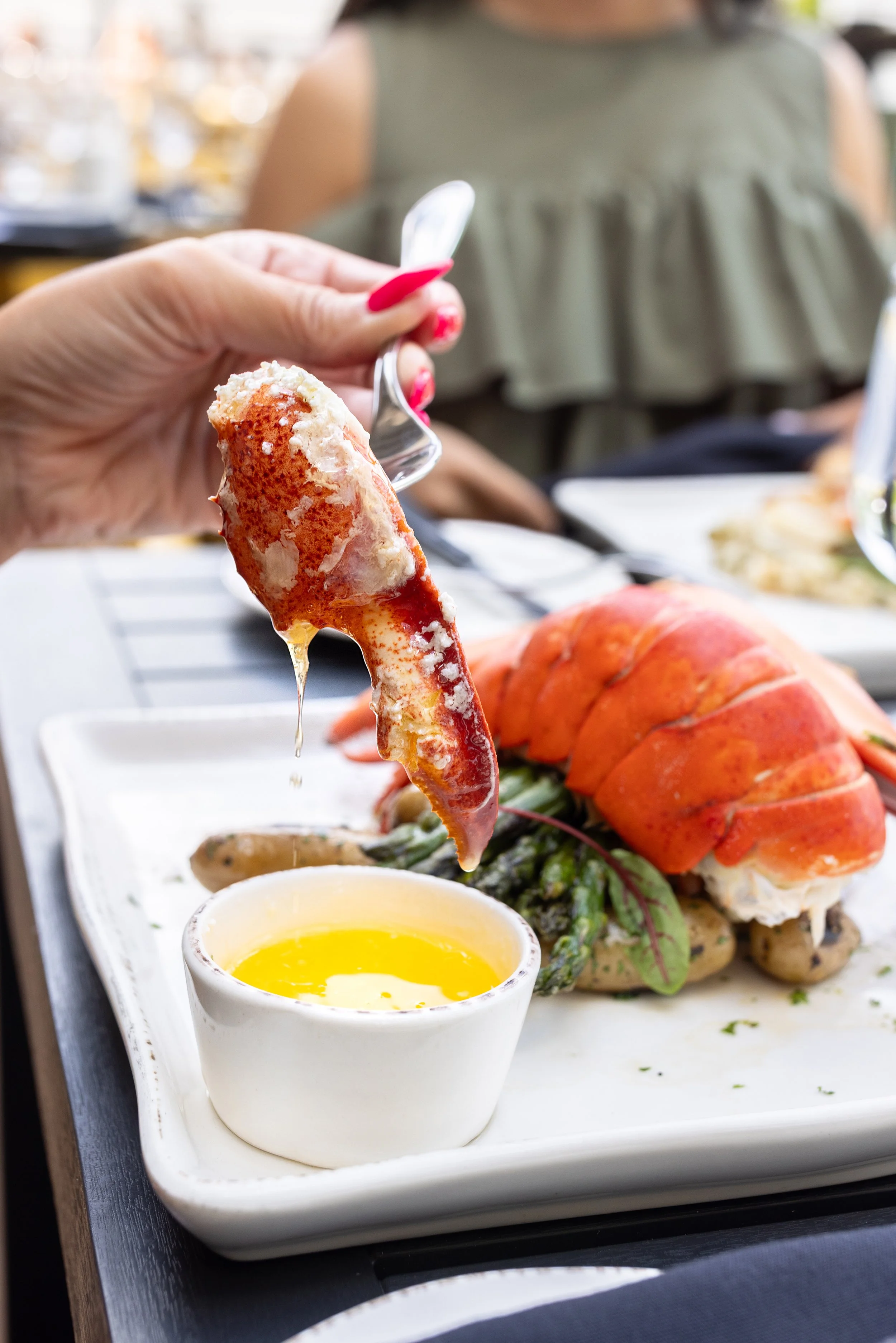 Maxwell's Steak & Seafood lobster dish. A person holding a lobster claw with melted butter dripping from it, with a lobster tail on a plate in the background.