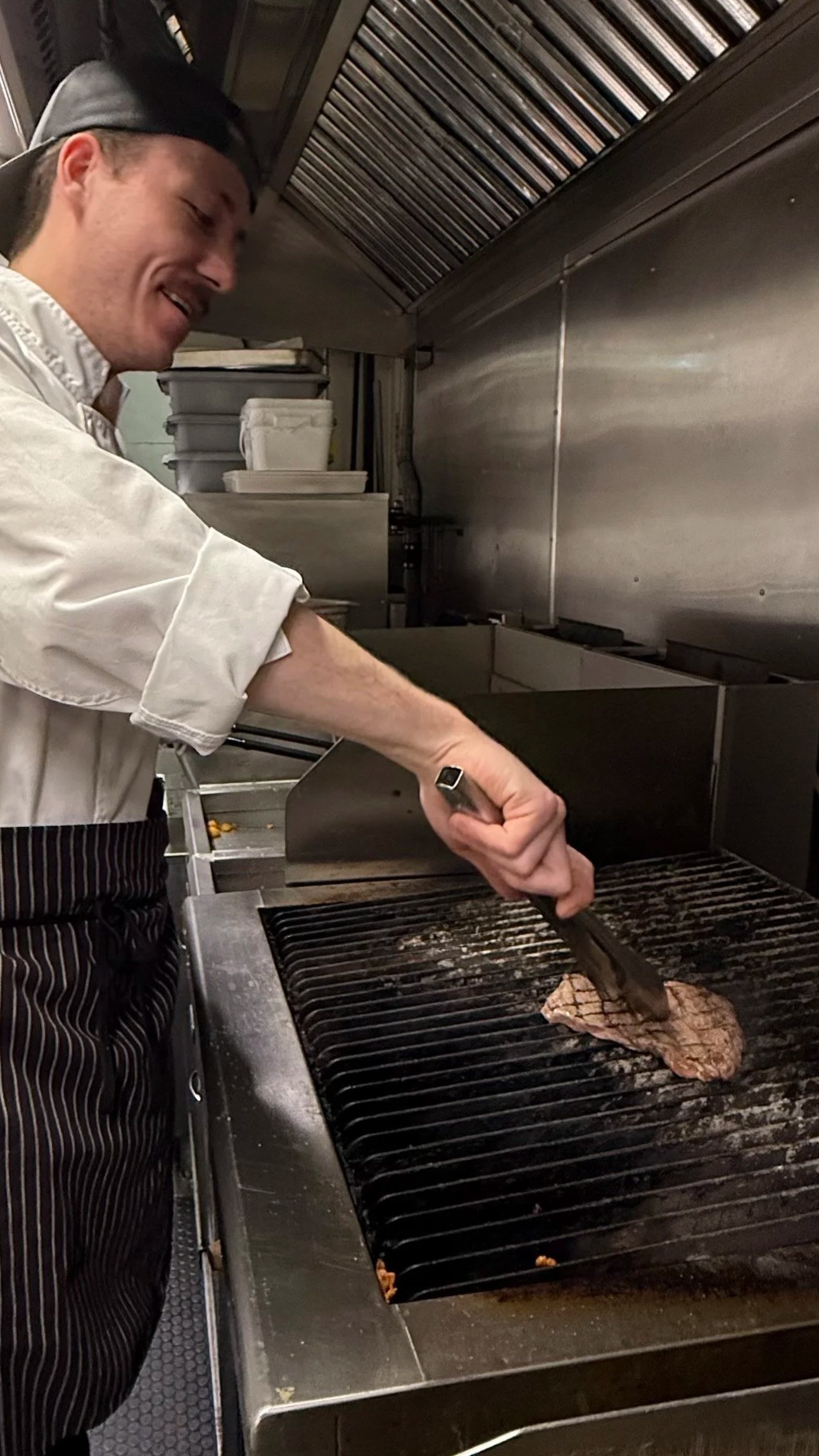 Chef Ryan wearing a striped apron and a white chef's coat is grilling a piece of steak on a commercial grill in Maxwell's kitchen.