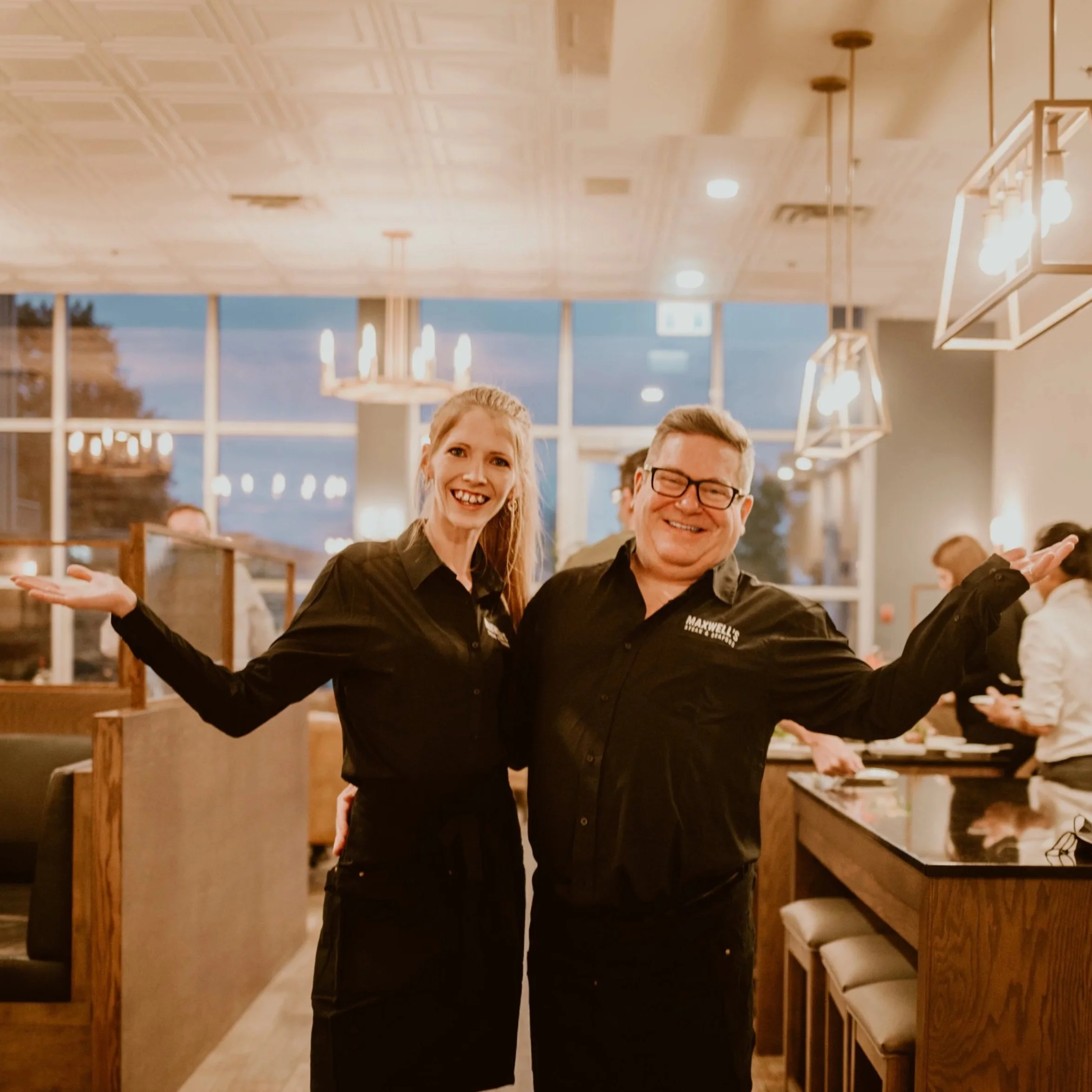 Two restaurant employees posing with arms outstretched inside the modern, well-lit dining area of Maxwell's Steak & Seafood with large windows in the background.