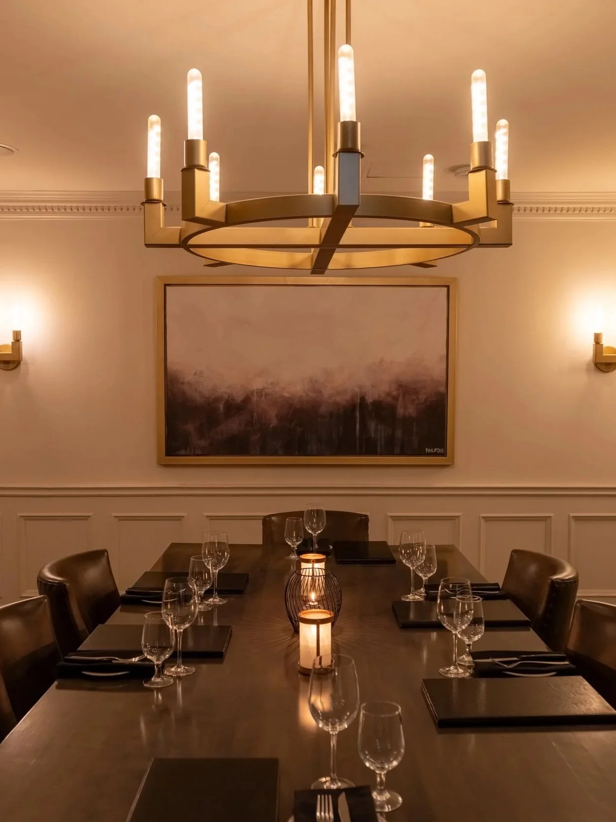 The elegant Websters dining room of Maxwell's with a long wooden table set with wine glasses and black napkins, candle holders with lit candles, a large chandelier hanging above, wall sconces, and a large framed abstract painting on the back wall.