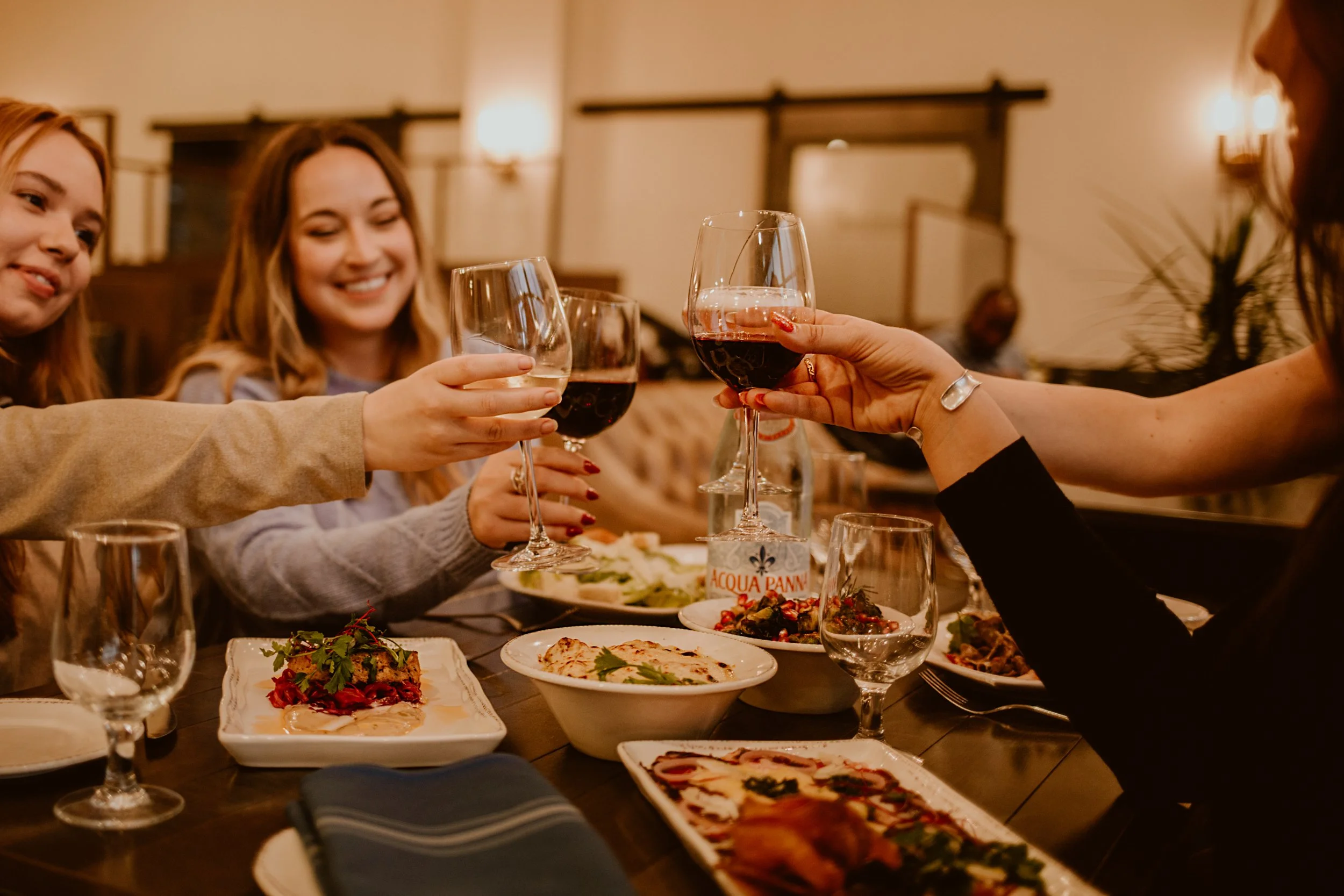 A group of women enjoying a meal together, raising glasses of red and white wine in a cheersing gesture over a table with various dishes in the warmly lit restaurant of Maxwell's Steak & Seafood.