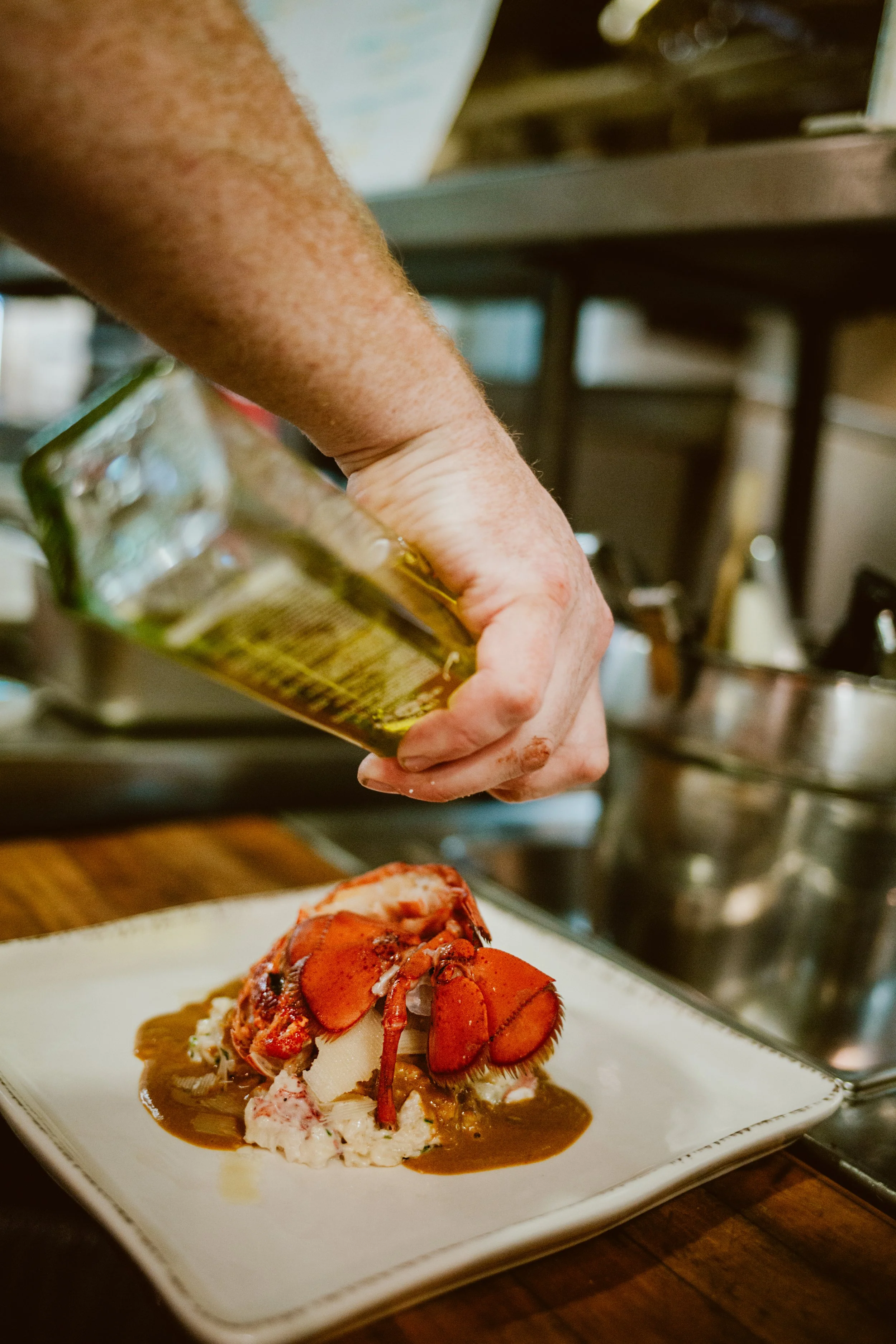 Maxwell's Steak & Seafood's chef's hand is pouring oil over a lobster dish on a white plate, garnished with sauce and other ingredients.