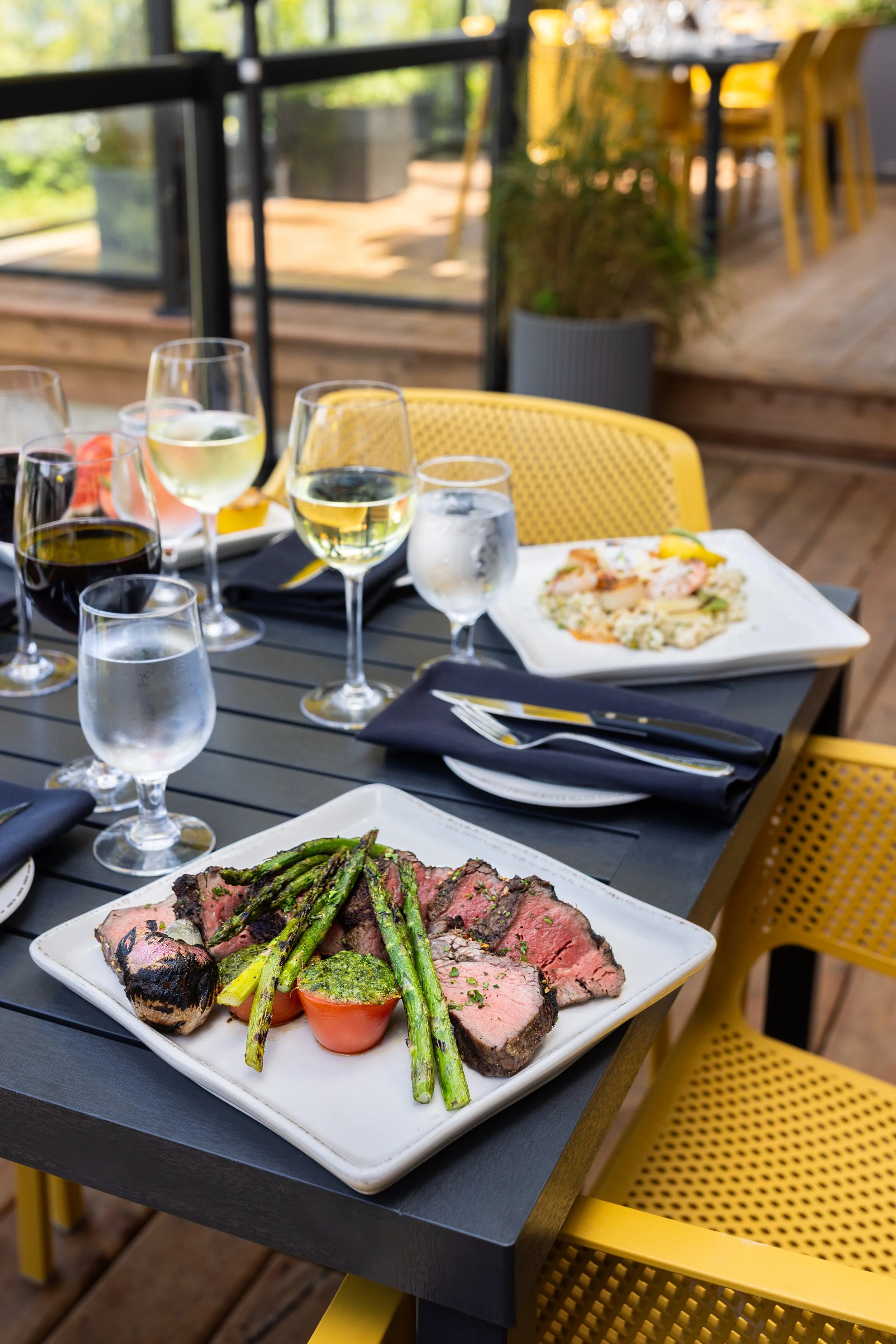 A dinner table set with various glasses of white and red wine, and plates of food including grilled steak with asparagus and a tomato, with seafood and vegetables, on a black patio table with yellow chairs outdoors at Maxwell's Steak & Seafood.