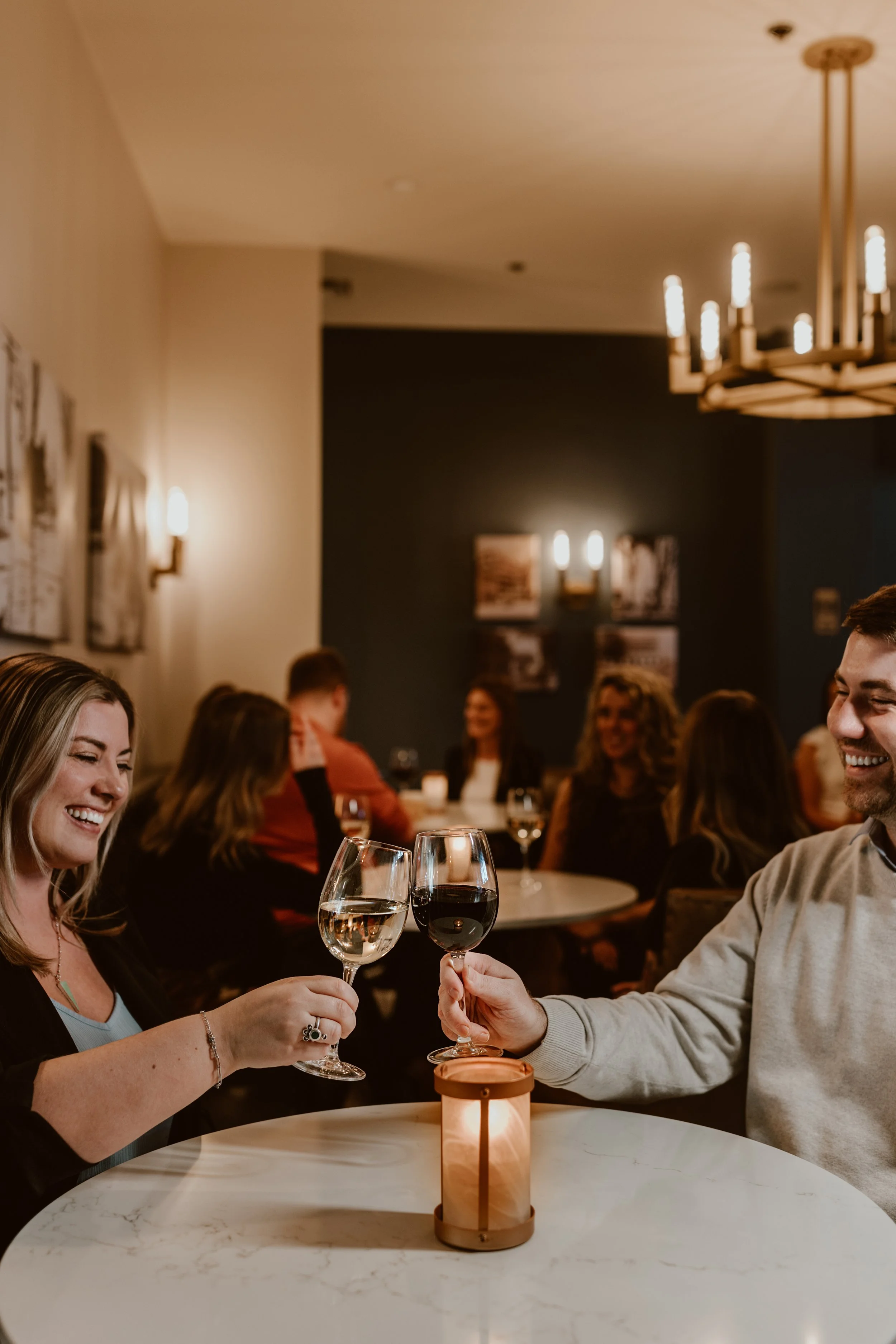 A couple clinking wine glasses, smiling at each other in the cozy Maxwell's Steak & Seafood lounge with dim lighting, other guests seated at tables in the background.