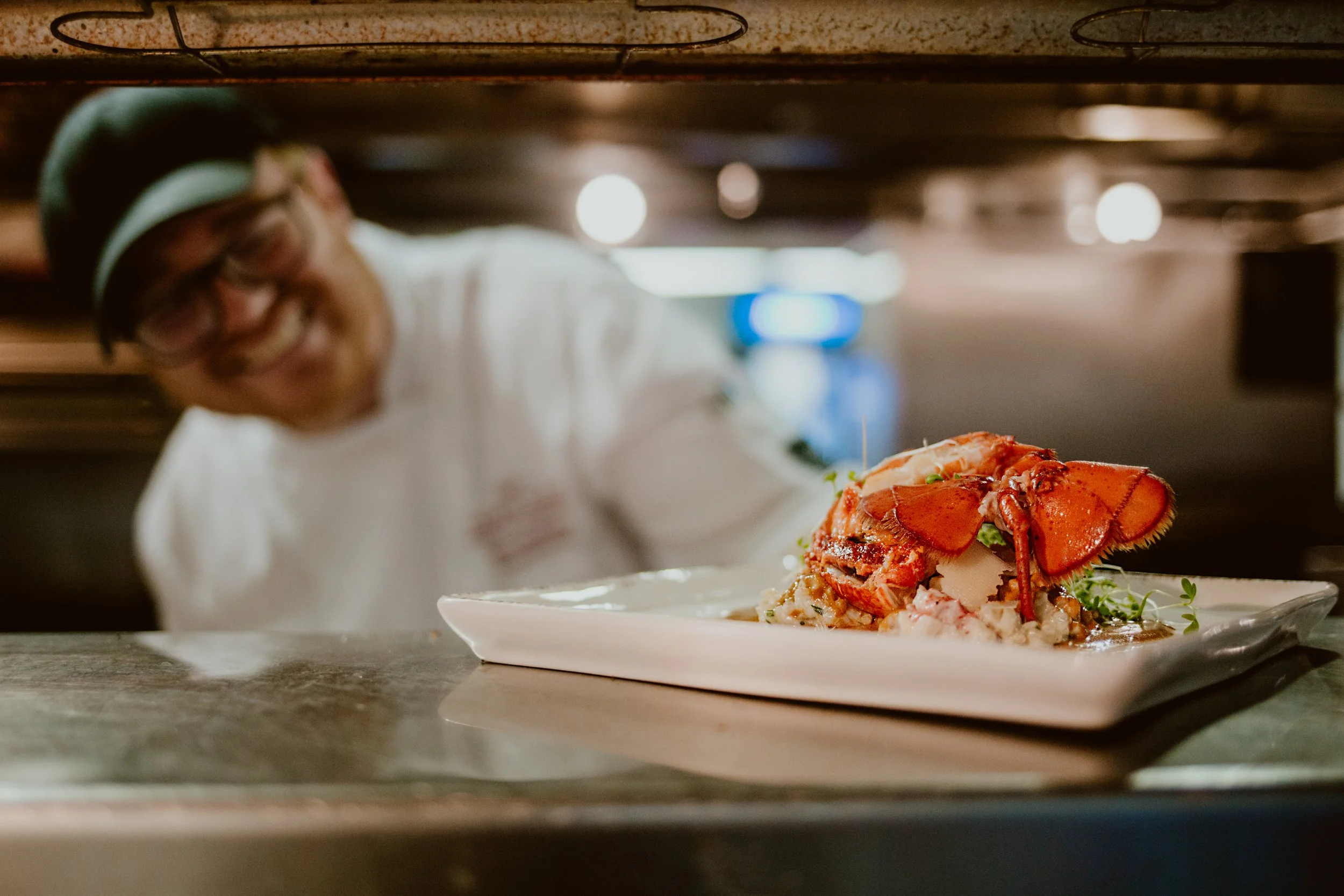 Chef Matt MacKenzie is seen smiling and leaning over a plate of seafood, including a lobster, on Maxwell's Steak & Seafood kitchen counter with a blurred background.