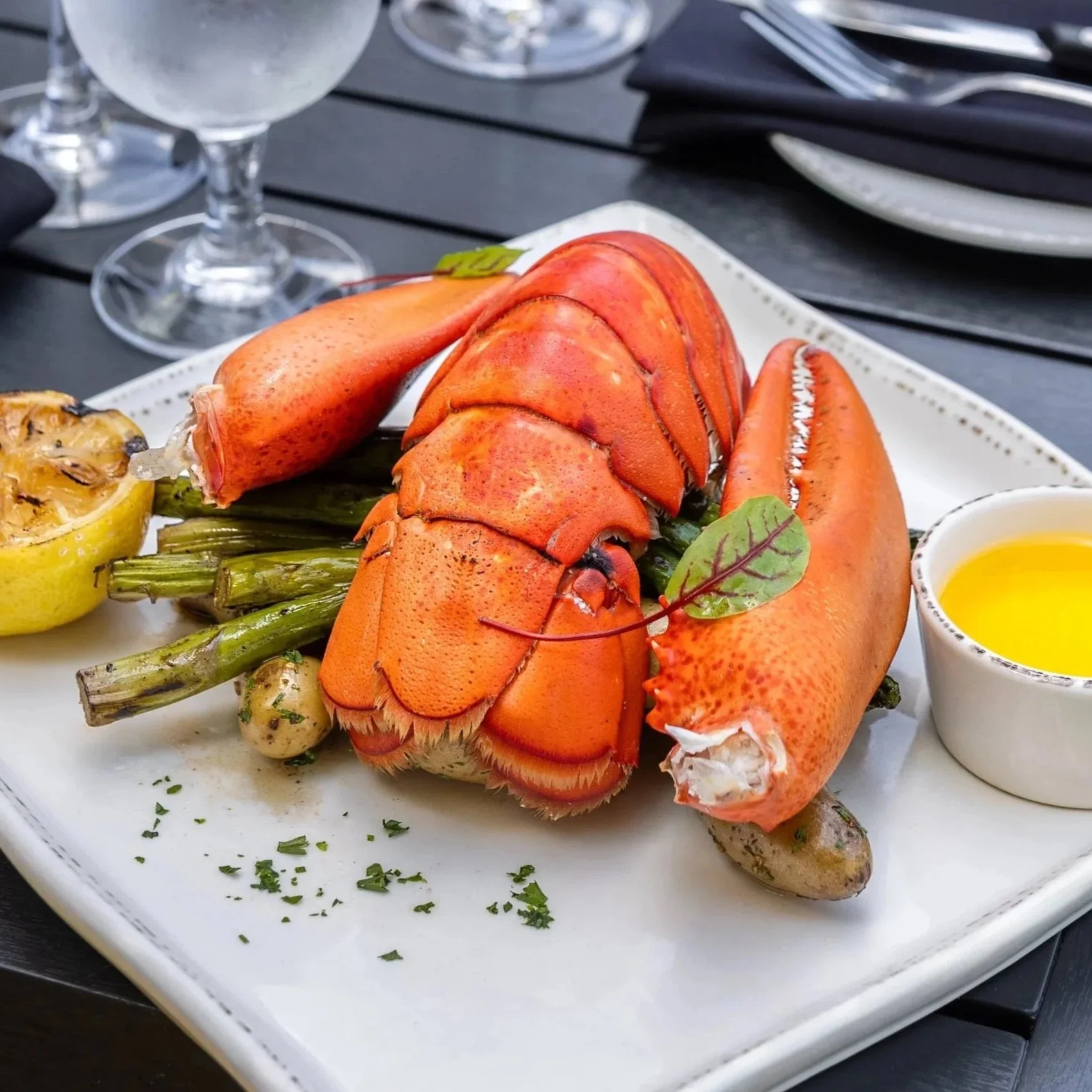 Cooked lobster tail served with grilled asparagus, a lemon half, and a small dish of melted butter on a white plate served on Maxwell's patio.