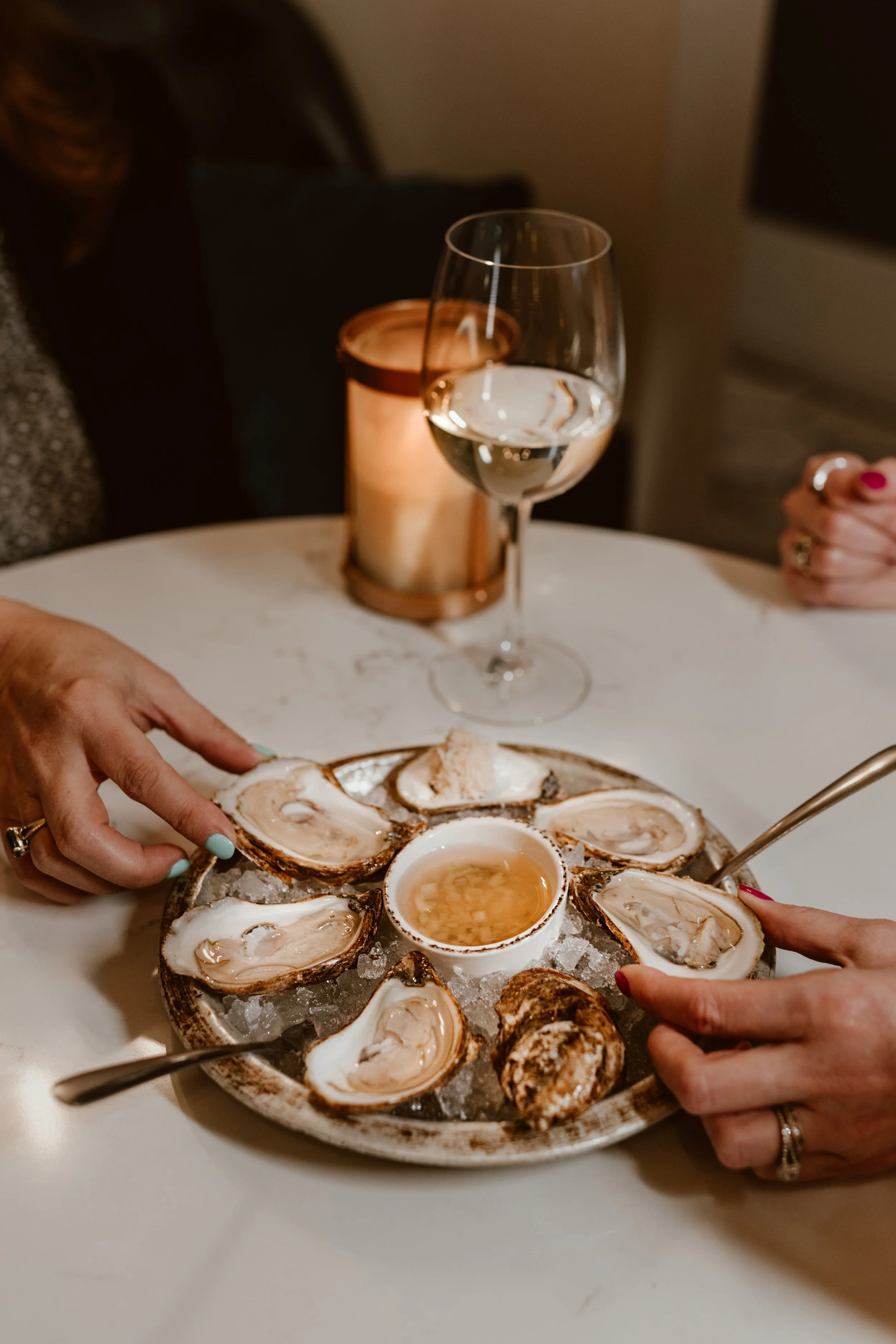 Plate of raw oysters on ice with a small cup of horseradish sauce in the centre, served at Maxwell's Steak & Seafood Restaurant with a glass of white wine, a candle, and part of a person's hand visible.