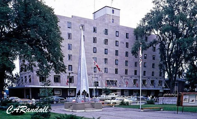 Fredericton city with a monument and three flagpoles, two of which have flags, in front of the Crowne Plaza Lord Beaverbrook Hotel, with parked cars and trees around.