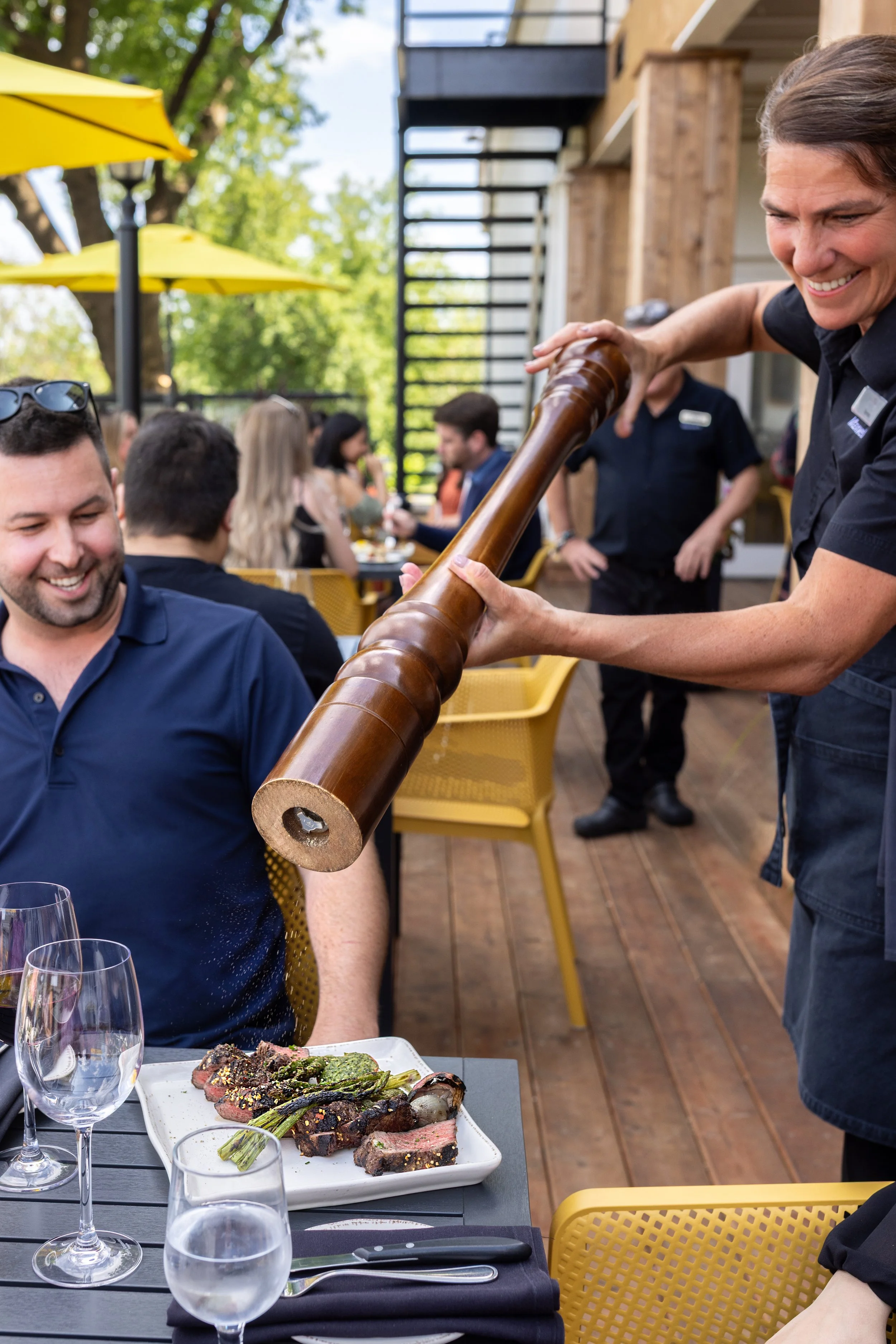 A server uses a large wooden pepper grinder to season a steak at the outdoor patio of Maxwell's Steak & Seafood with yellow umbrellas and green trees.