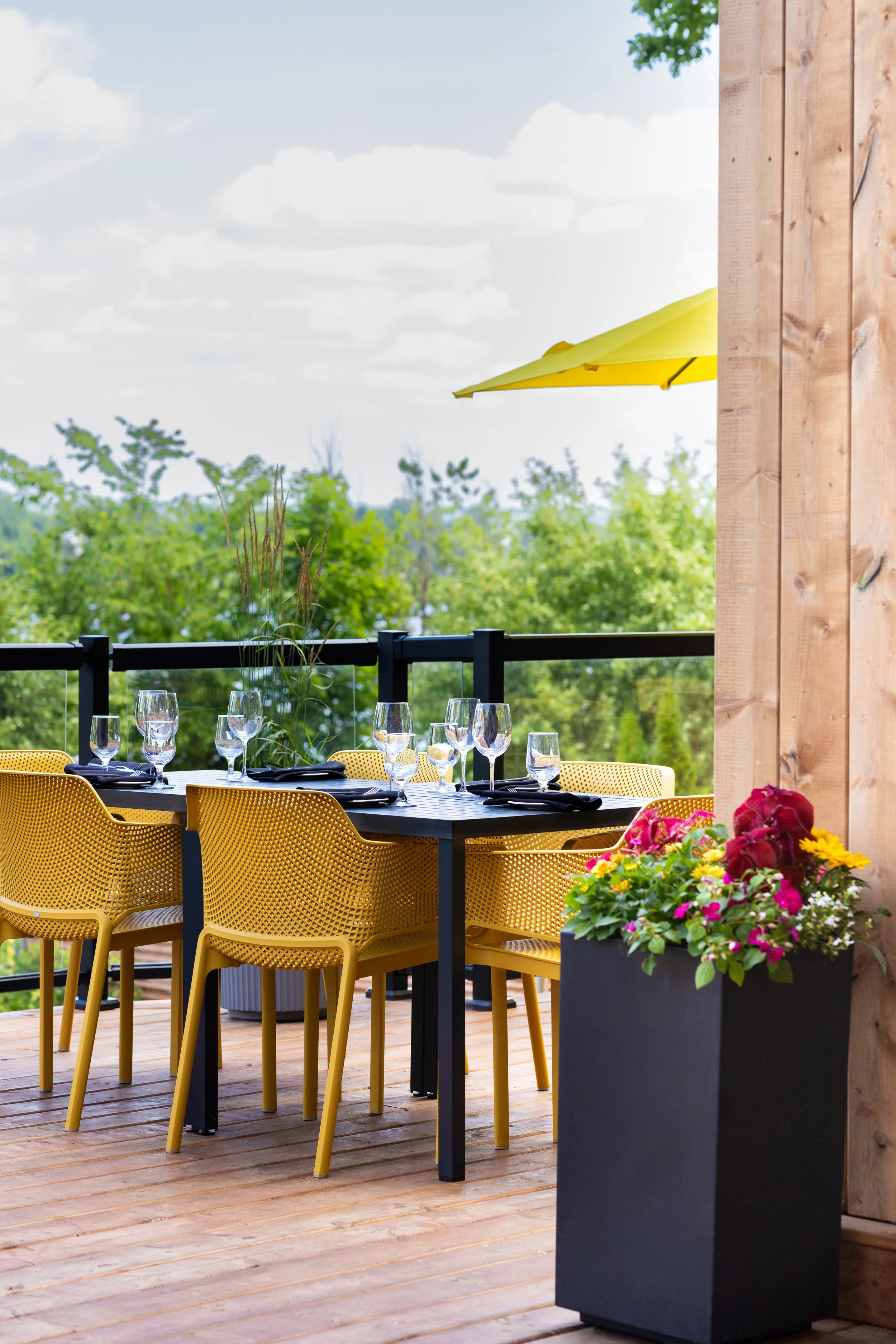 The outdoor dining area of Maxwell's Steak & Seafood, black table set with glasses and black napkins, surrounded by yellow chairs on a wooden deck, overlooking green trees under a blue sky with clouds.
