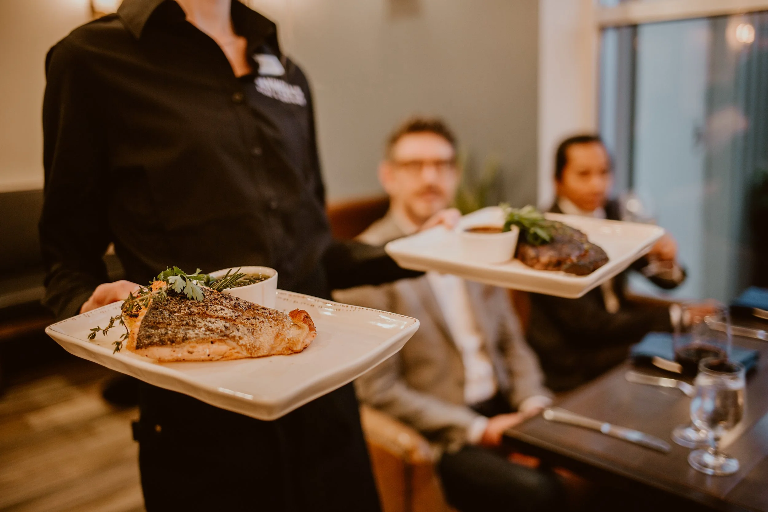 Server holding two plates with cooked fish and a steak, herbs, and sauce in Maxwell's Steak & Seafood restaurant dining area with three seated guests in the background.