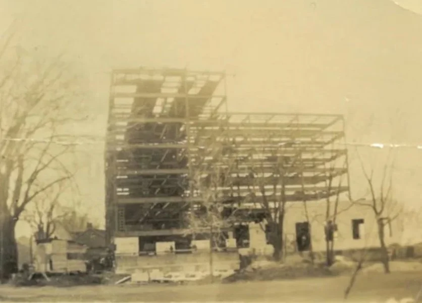 Black and white photo of the Crowne Plaza Fredericton - Lord Beaverbrook hotel under construction, with steel framework visible, surrounded by leafless trees and some older small buildings.