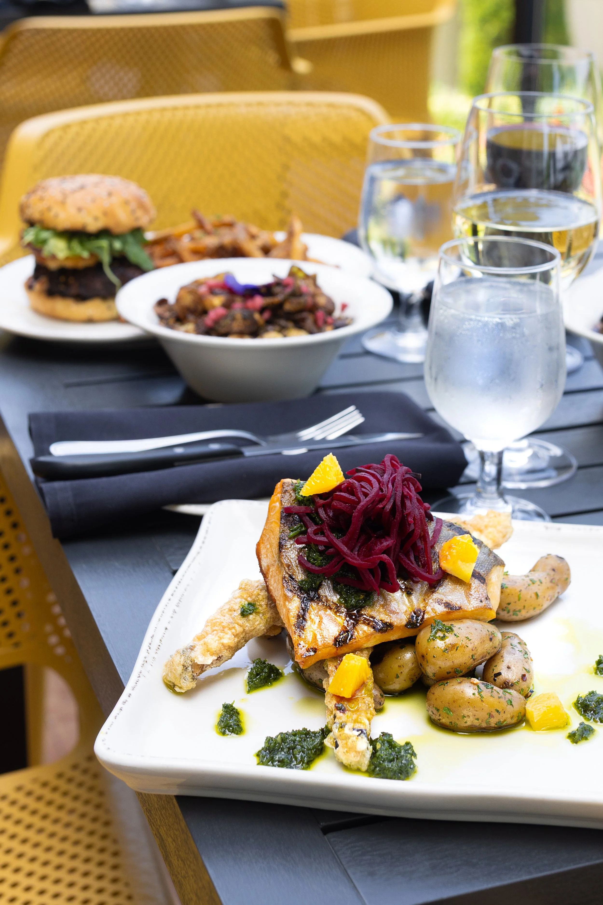 Dining table with a plated fish dish topped with beets and yellow garnish, several glasses of water and wine, a burger with fries, and a bowl of food in the background, with yellow chairs nearby served at Maxwell's Steak & Seafood patio.