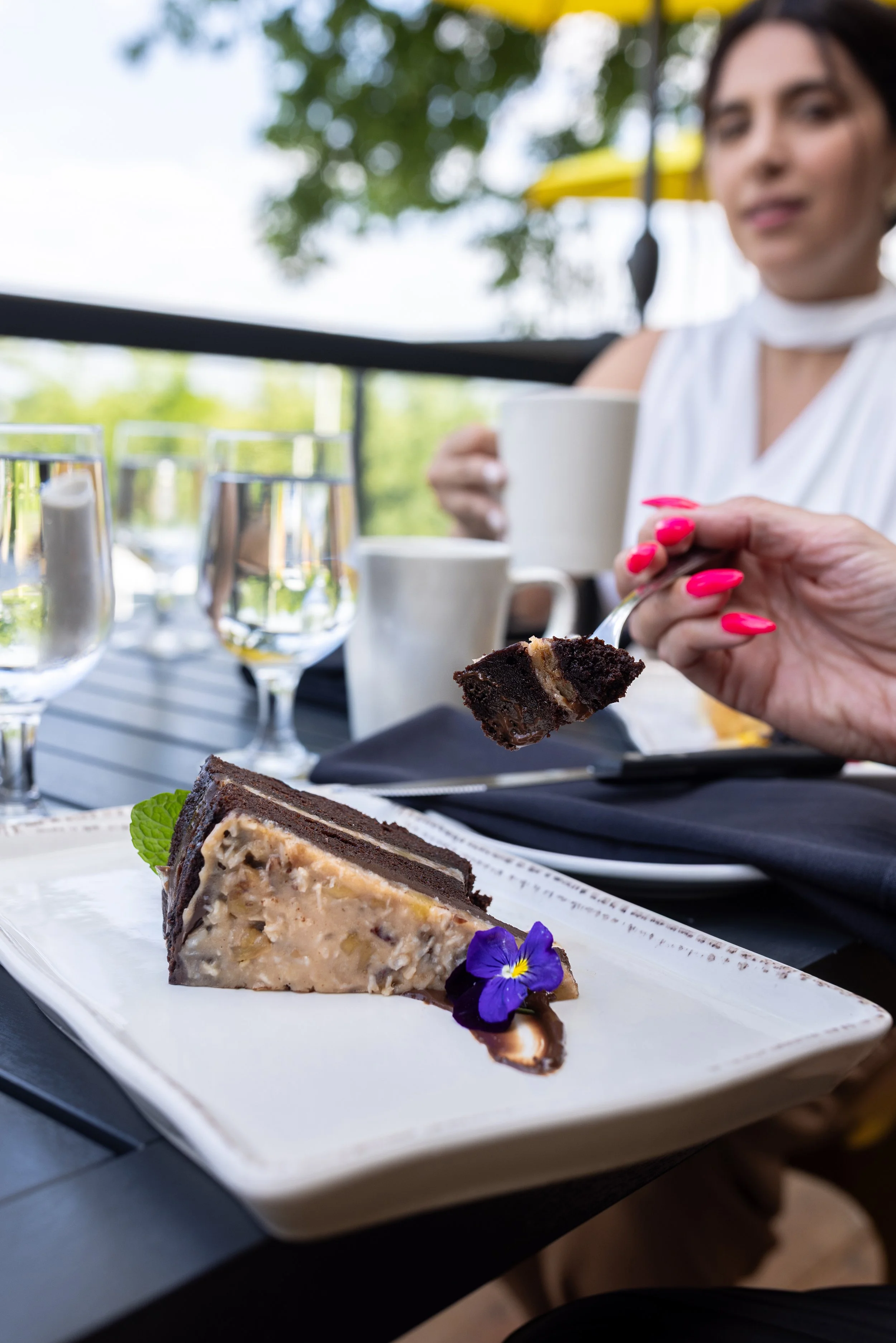 Close-up of a dessert plate with a slice of Maxwell's Steak & Seafood chocolate cake garnished with an edible flower, on a black table with glasses and cups in the background, at an outdoor dining setting.