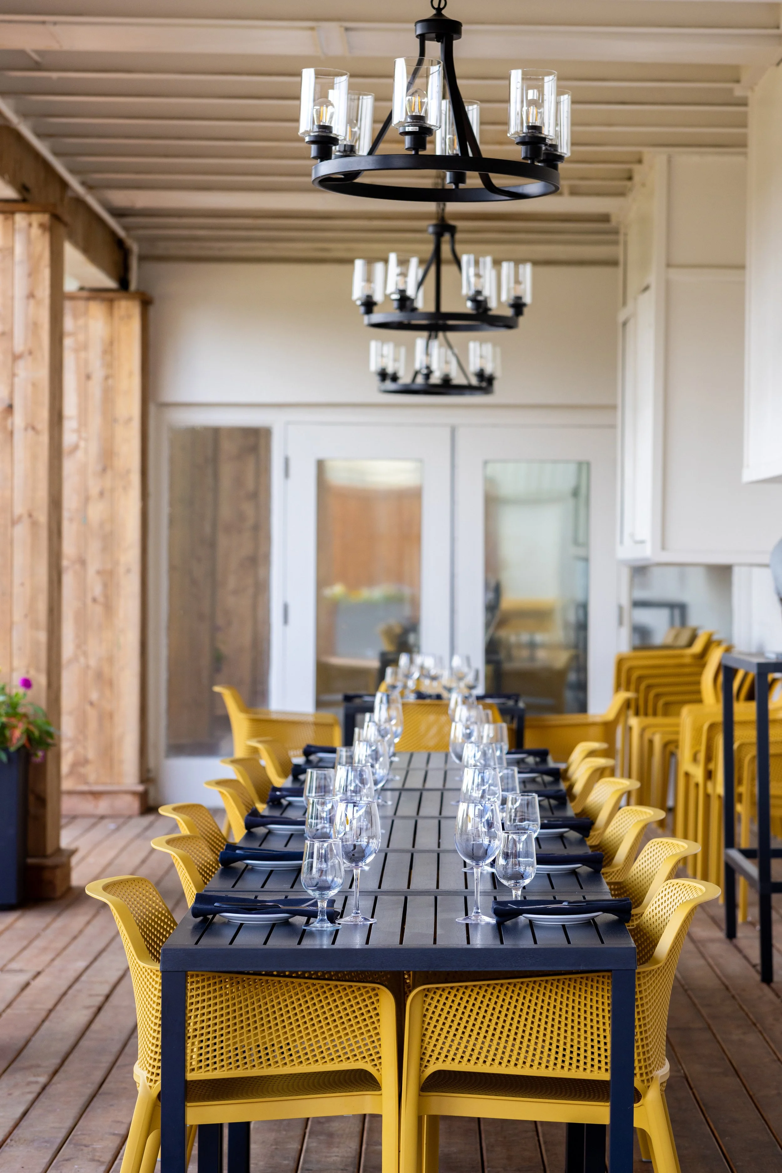 A dining table set for a meal on Maxwell's Steak & Seafood patio with plates, wine glasses, and black napkins, surrounded by yellow chairs, on a wooden deck with chandeliers hanging above.